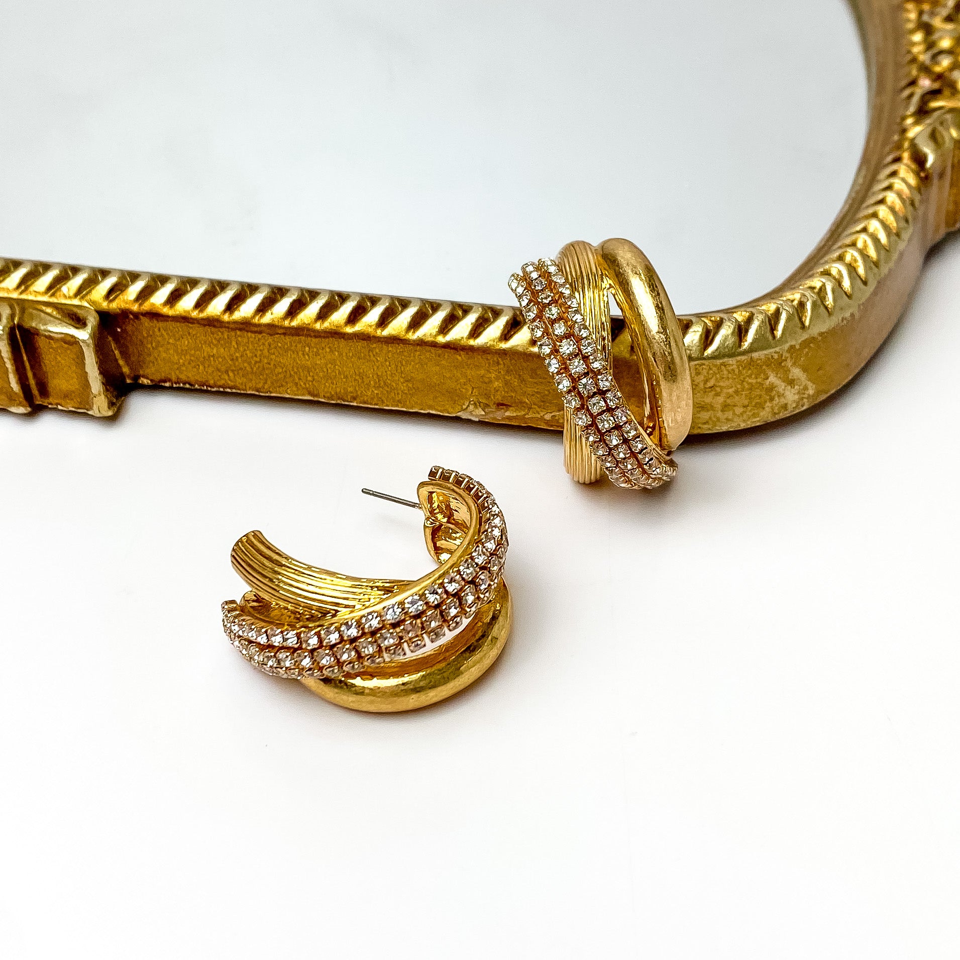 Three banded gold hoop earrings that are intertwined together. There is a smooth band, a striped band, and a crystal band. These earrings are pictured on a white background with one hoop leaning on a gold mirror. 