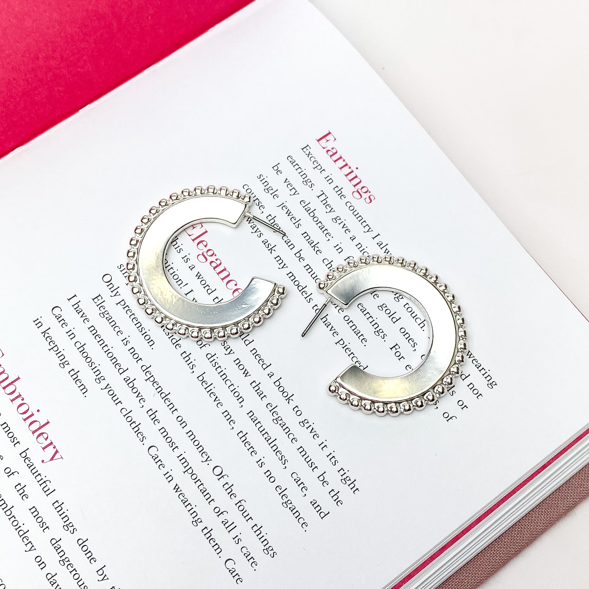 Smooth, silver hoop earrings with a beaded edge. These earrings are pictured on an open book on a white background. 