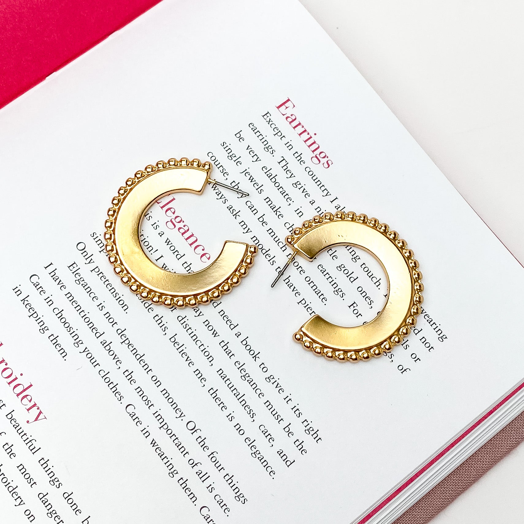 Smooth, gold hoop earrings with a beaded edge. These earrings are pictured on an open book on a white background. 