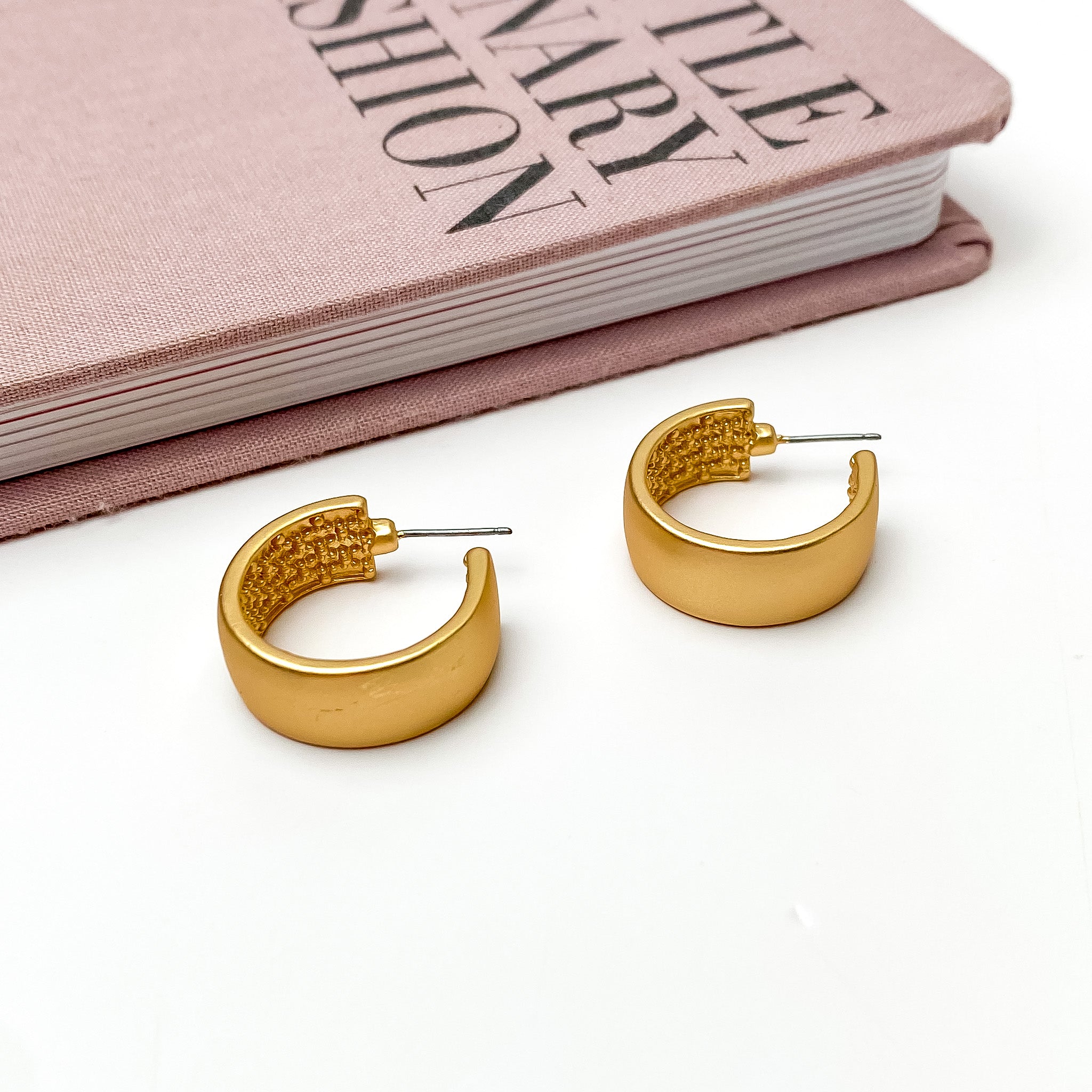 Gold Tone Hoop Earrings With a Textured Inside. Pictured on a white background with a pink book above the earrings.