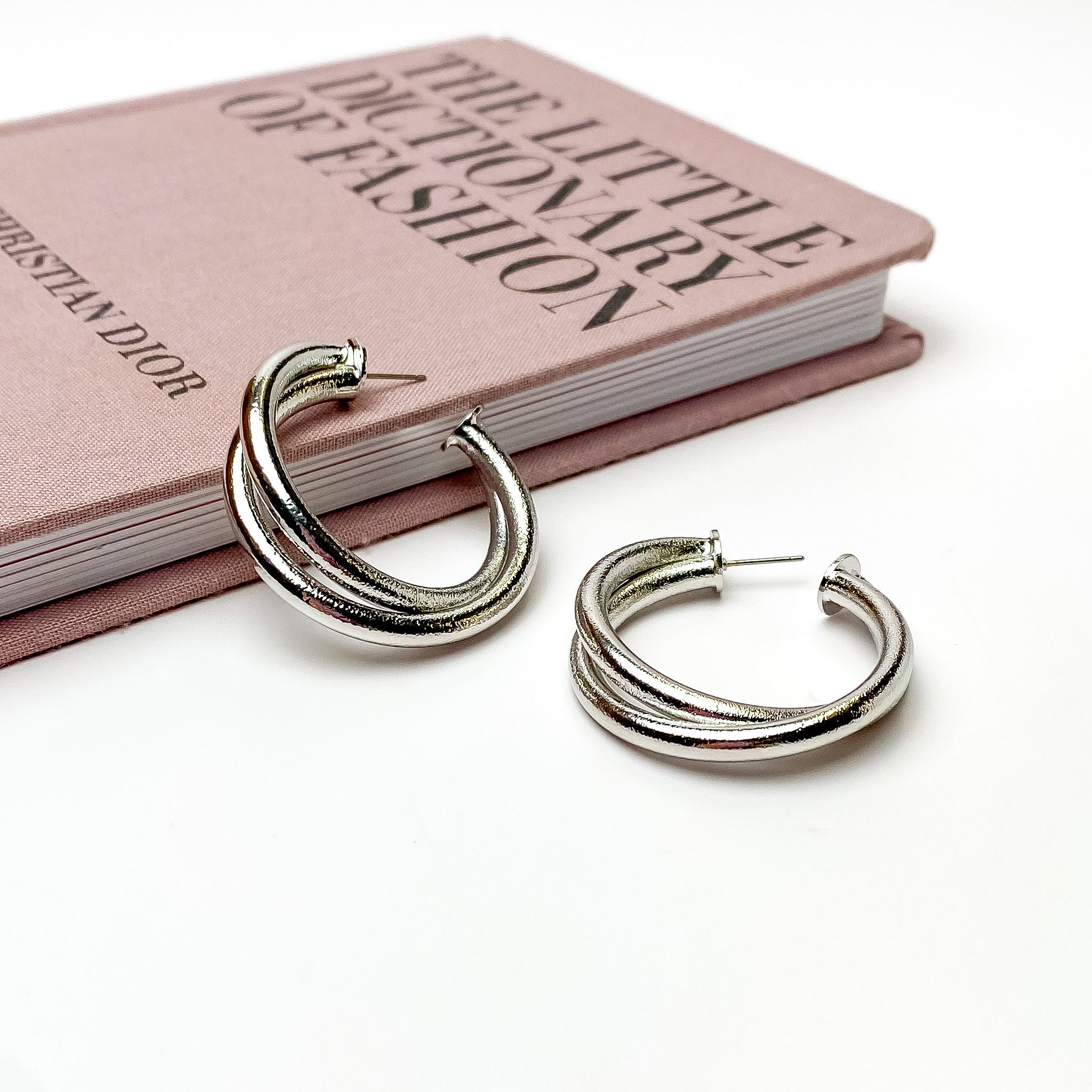 Silver, textured twisted hoop earrings. These earrings are pictured on a white background with one hoop is laying against a mauve colored book. 