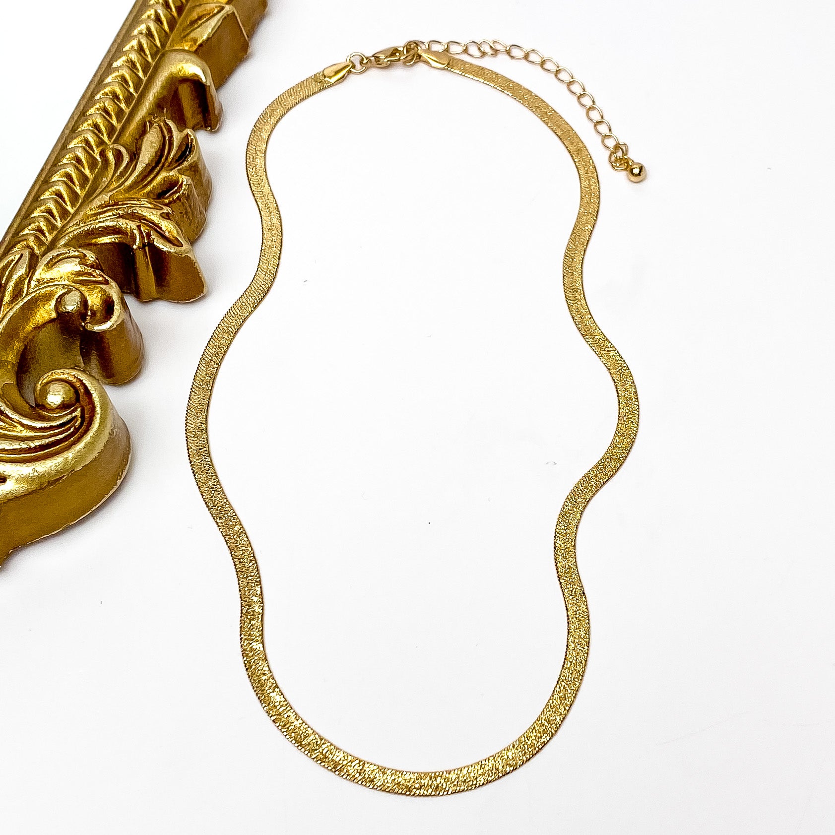 Gold herringbone chain necklace with a gold necklace adjuster. This necklace is pictured on a white background with a gold mirror on the left side. 