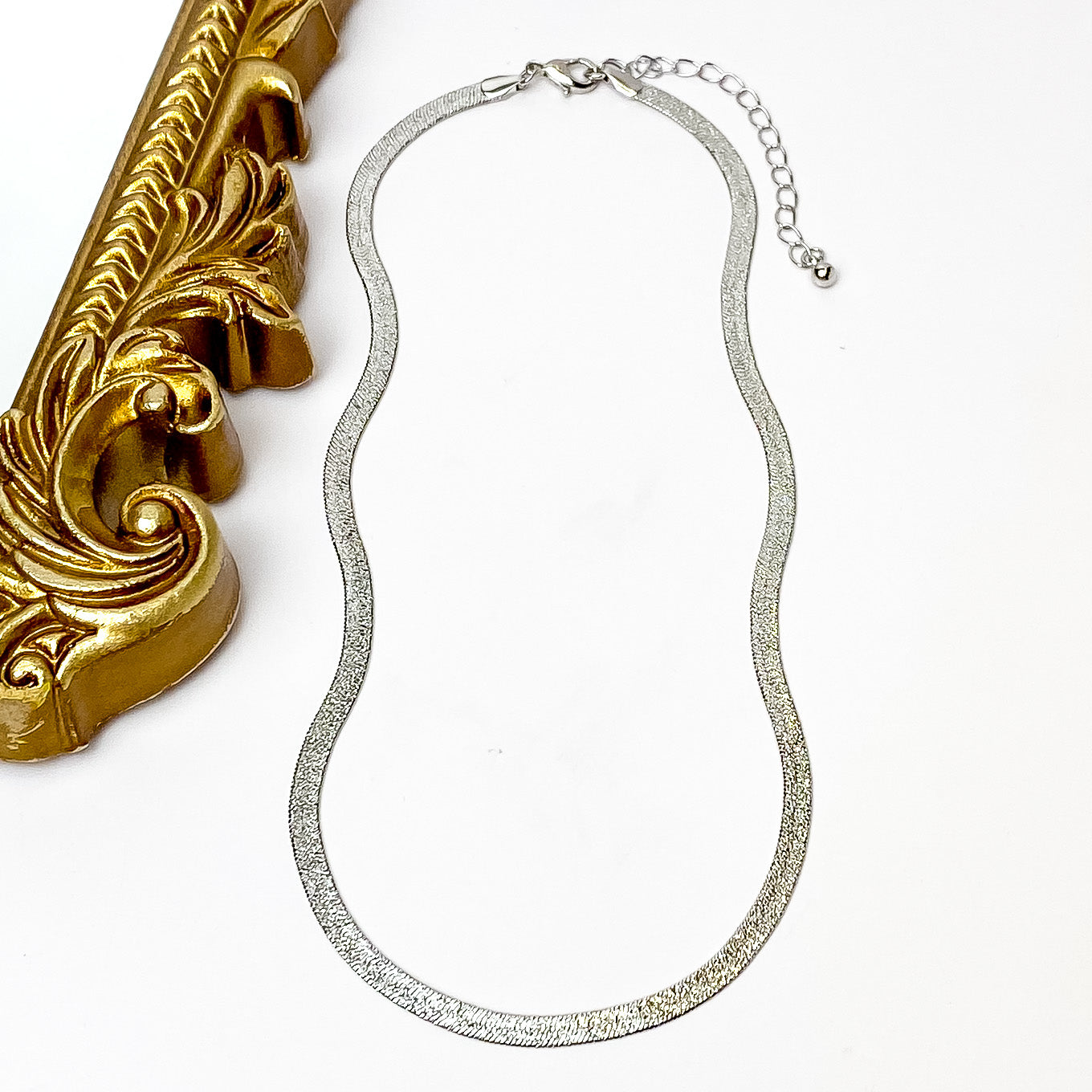 Silver herringbone chain necklace with a silver necklace adjuster. This necklace is pictured on a white background with a gold mirror on the left side. 