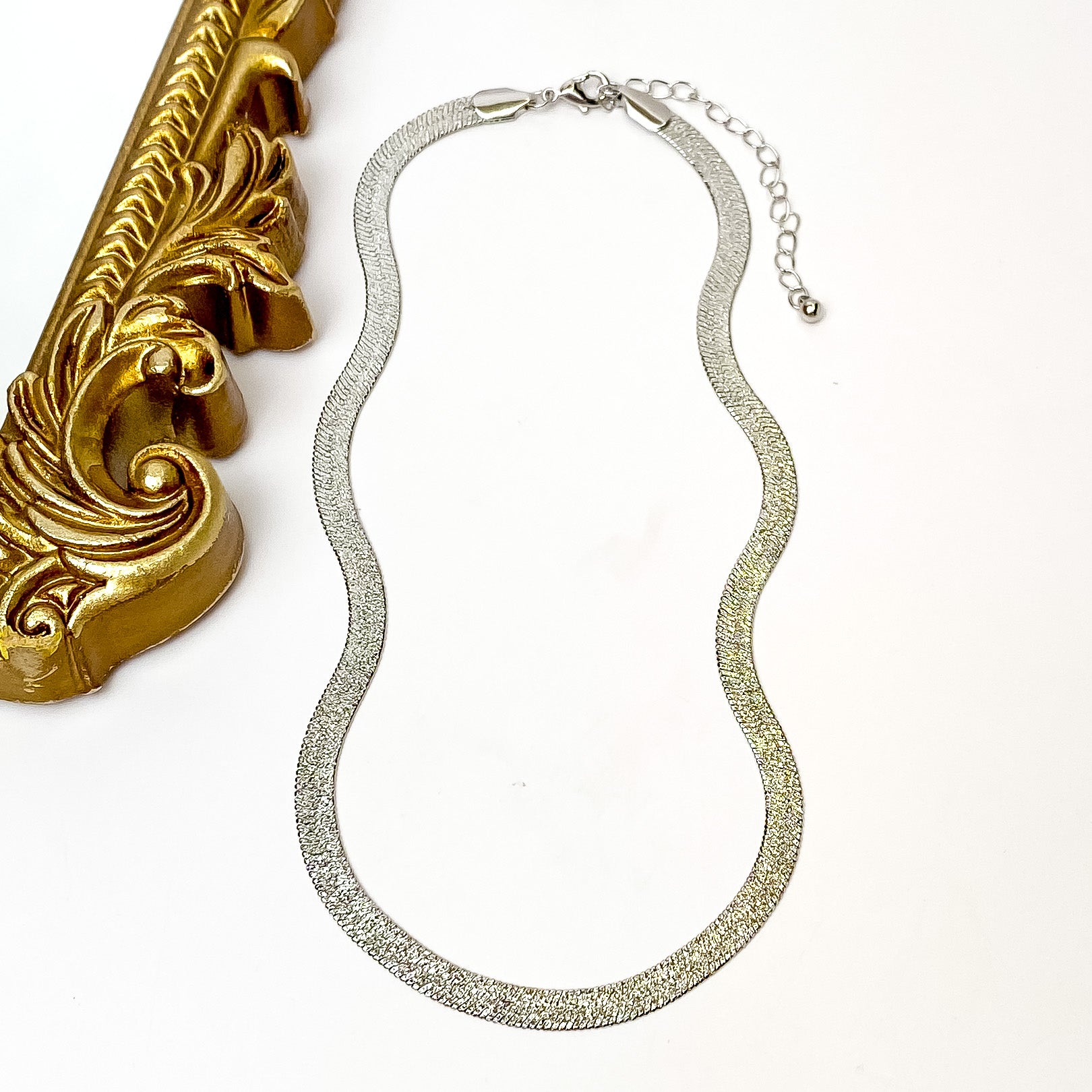 Silver herringbone chain necklace with a silver necklace adjuster. This necklace is pictured on a white background with a gold mirror on the left side. 