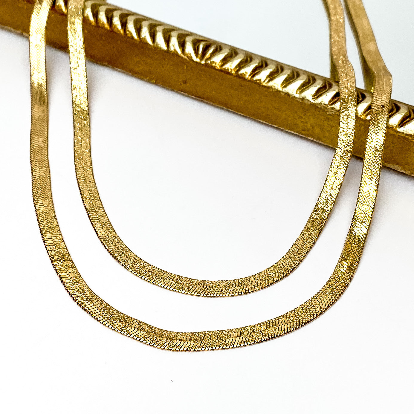 Double layered gold herringbone chain necklace. The longer strand has star imprints throughout the chain. This necklace is pictured partially laying on a gold mirror on a white background. 