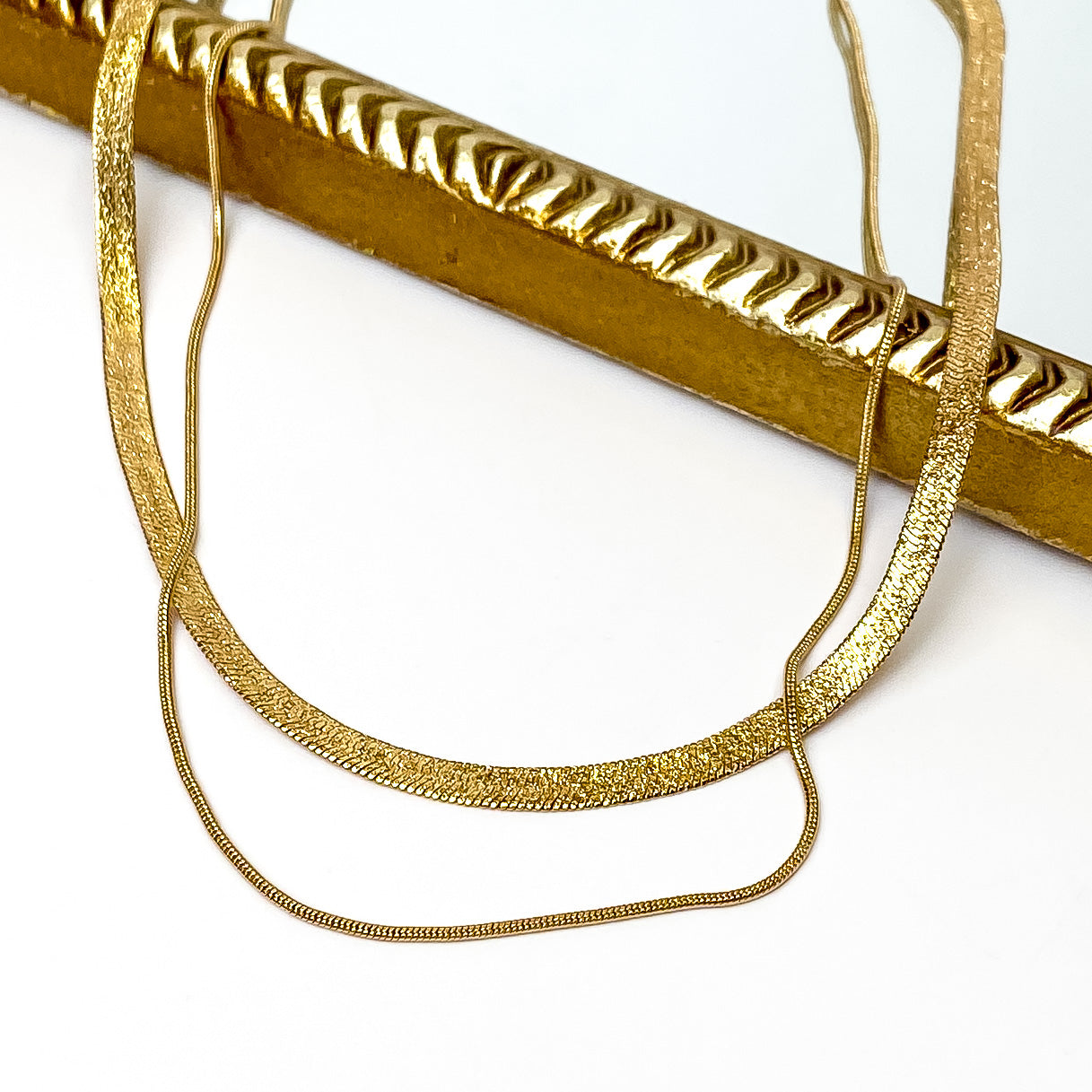 Gold herringbone chain and thin rope chain layered necklace. This necklace is pictured partially laying on a gold mirror on a white background. 