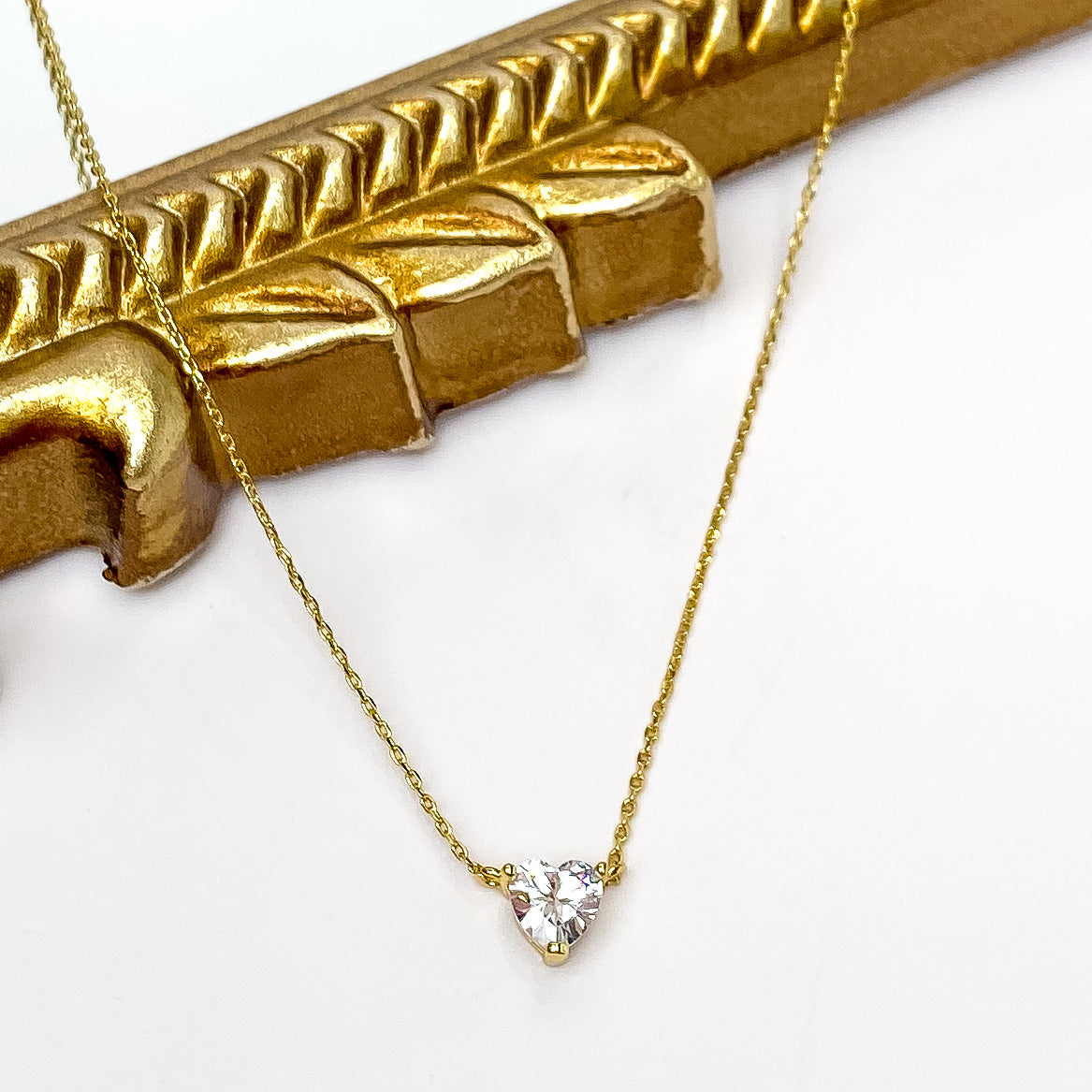 Gold chain necklace with a clear crystal heart pendant. This necklace is pictured partially laying on a gold mirror on a white background. 