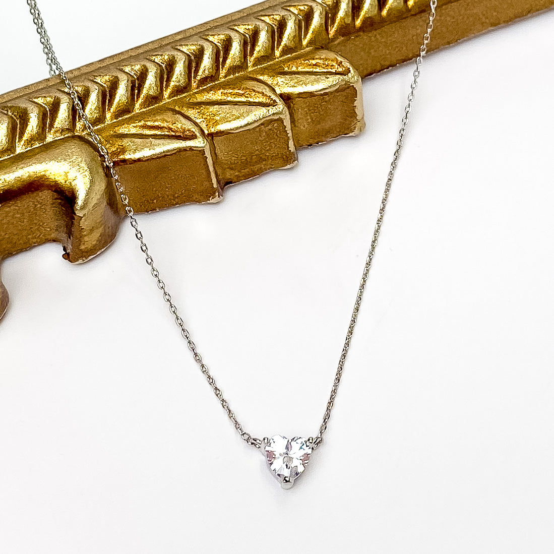 Silver chain necklace with a clear crystal heart pendant. This necklace is pictured partially laying on a gold mirror on a white background. 