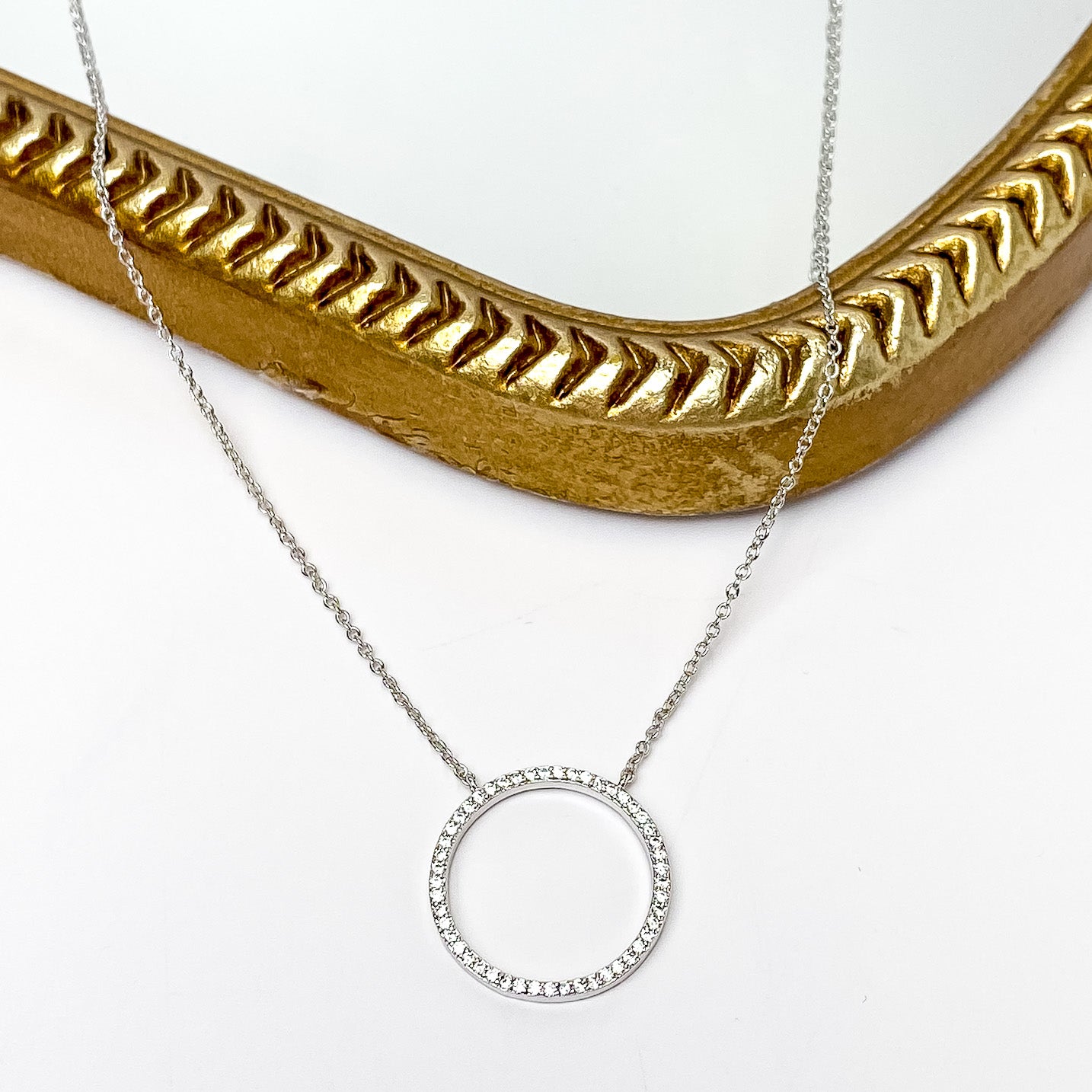 Silver chain necklace with an open circle pendant that has a cz crystal inlay. This necklace is pictured partially laying on a gold mirror on a white background. 