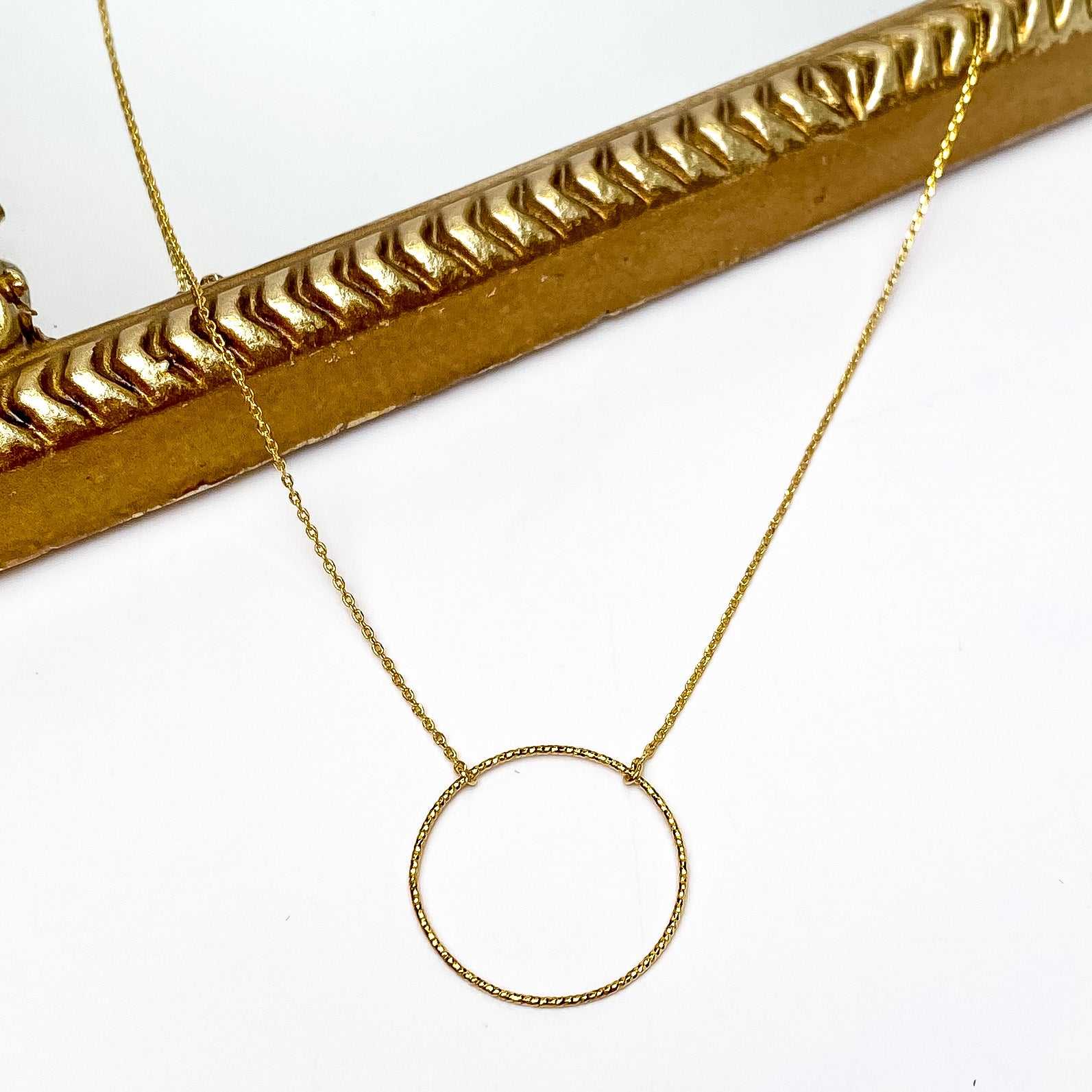 Dainty gold chain necklace with a hammered, open circle pendant. This necklace is pictured partially laying on a gold mirror on a white background. 