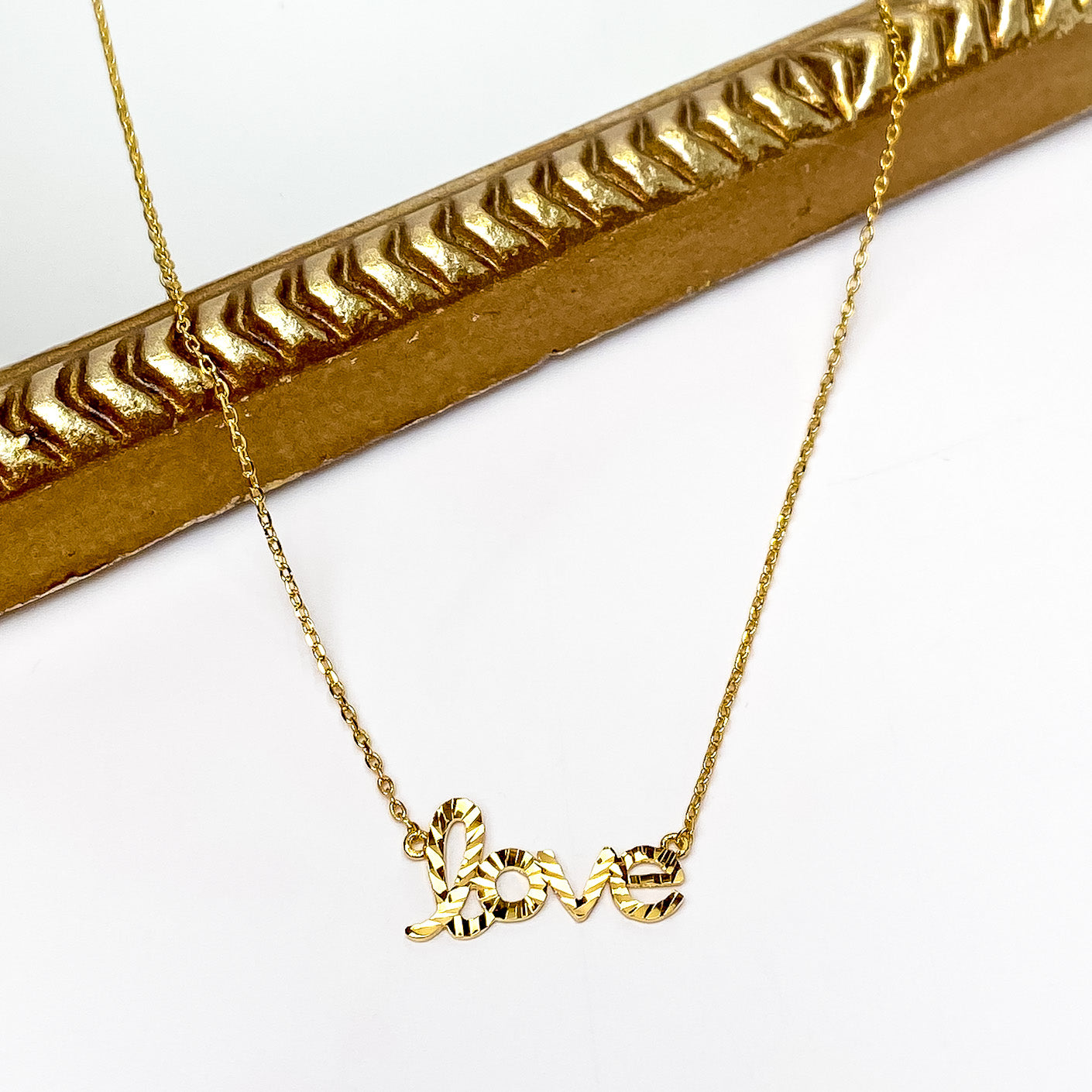 Gold chain necklace with a pedant that spells out "love". This necklace is pictured partially laying on a gold mirror on a white background. 
