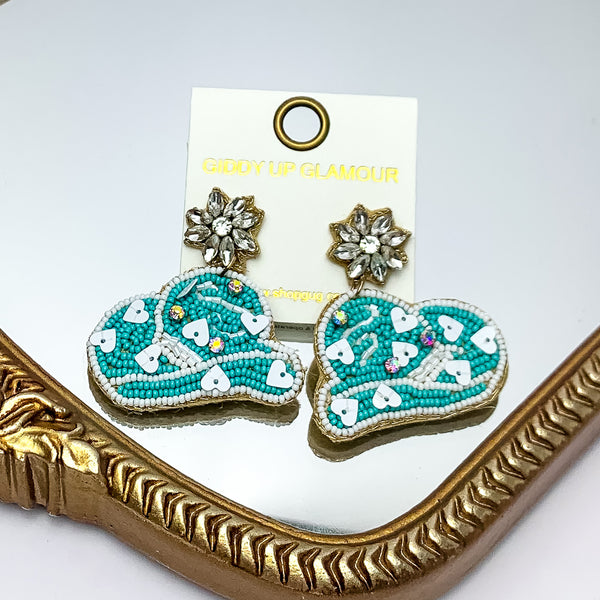 White and turquoise blue beaded cowboy hat earrings. Crystal flower posts. Pictured on a mirror with a gold trim. 