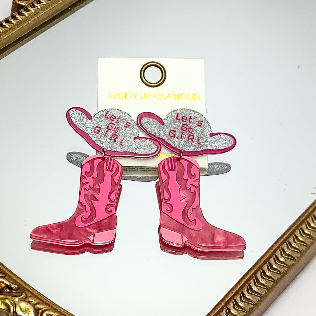 Let's Go Girl Cowboy Boot Earrings With Cowboy Hat Posts in Pink. Pictured on a mirror with a gold trim.