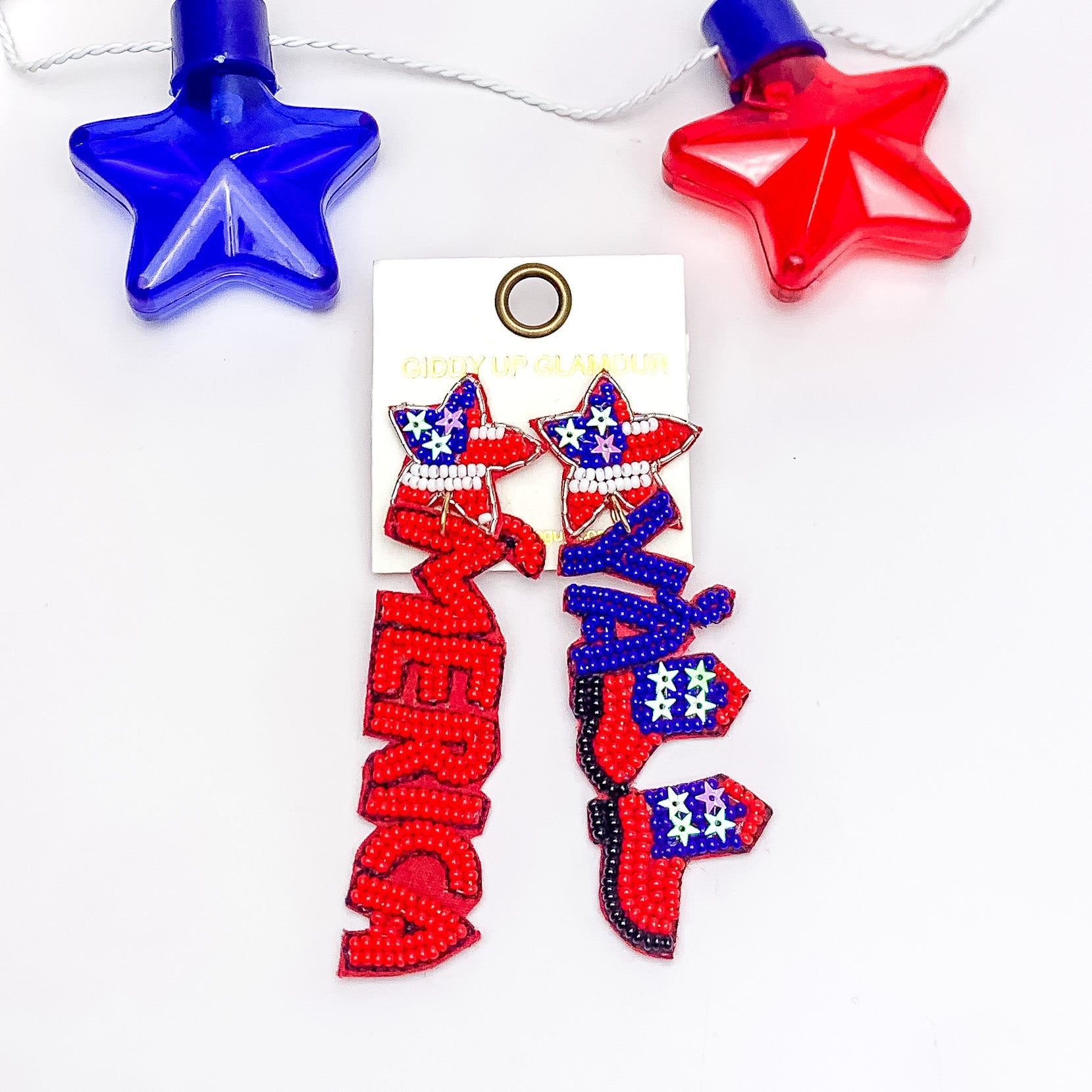Star shaped post earrings with white and red stripes and a blue part with star sequins. One earrings has a red pendant that spells ot "'MERICA" and the other one has a pendant that spells out "Y'ALL." The y and a include blue beads while the two Ls are in the shape of boots. These earrings are pictured on a white background. 