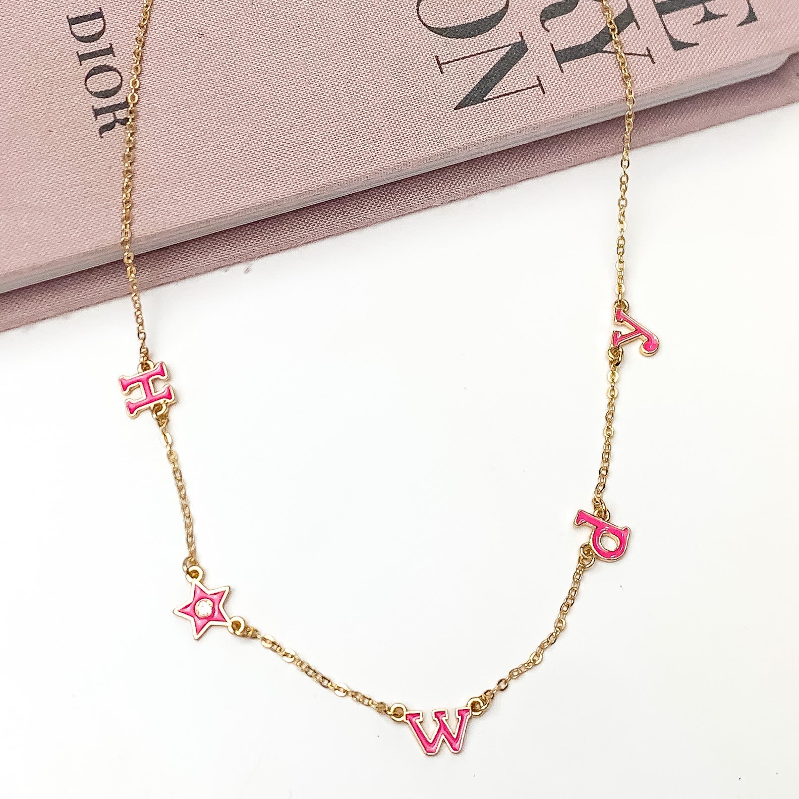 Howdy Gold Tone Chain Necklace in Dark Pink. Pictured on a white background with the top of the necklace laying on a book.