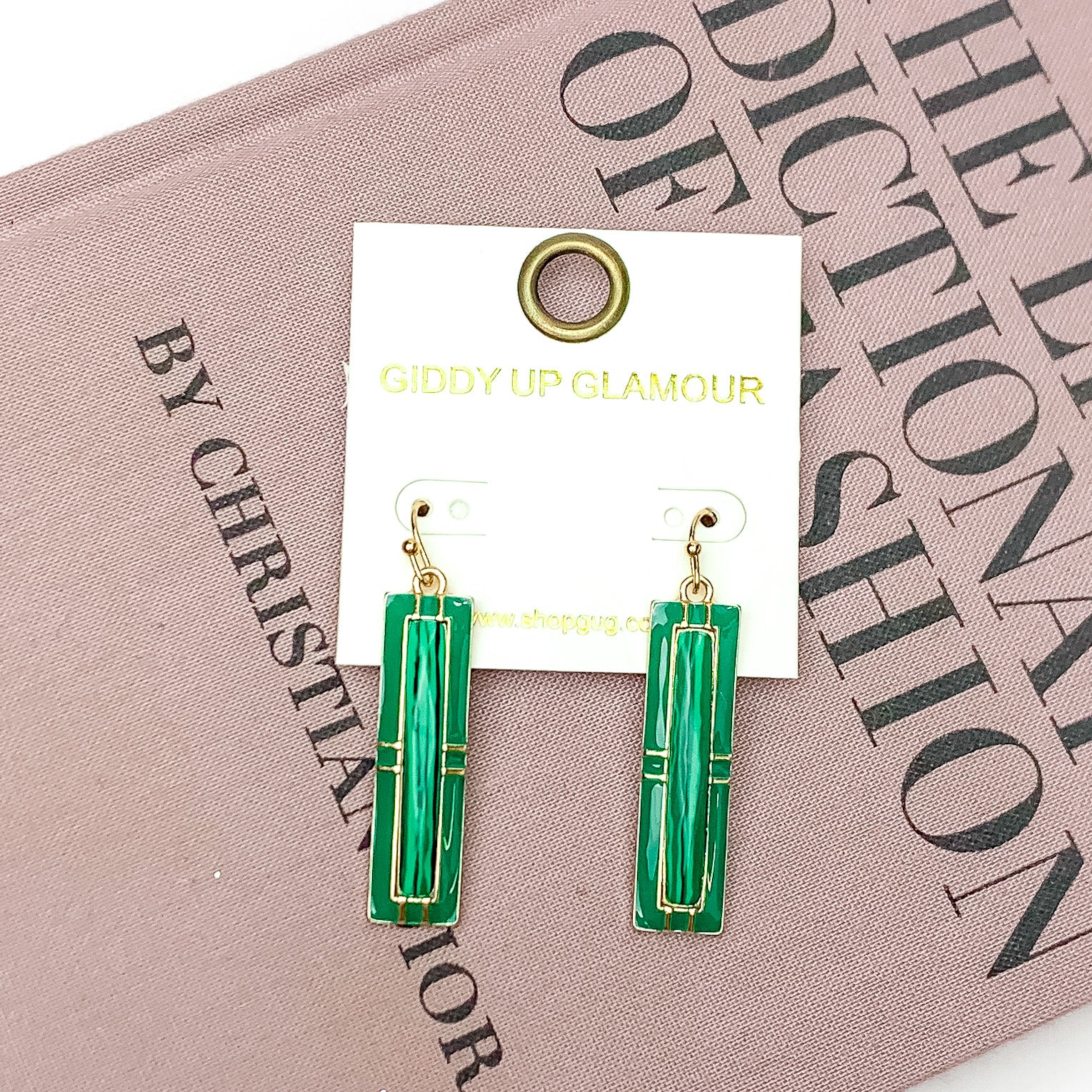 Everyday Gold Tone Rectangular Pendant Earrings in Kelly Green. Pictured on a white background with the earrings laying on a pink book. 