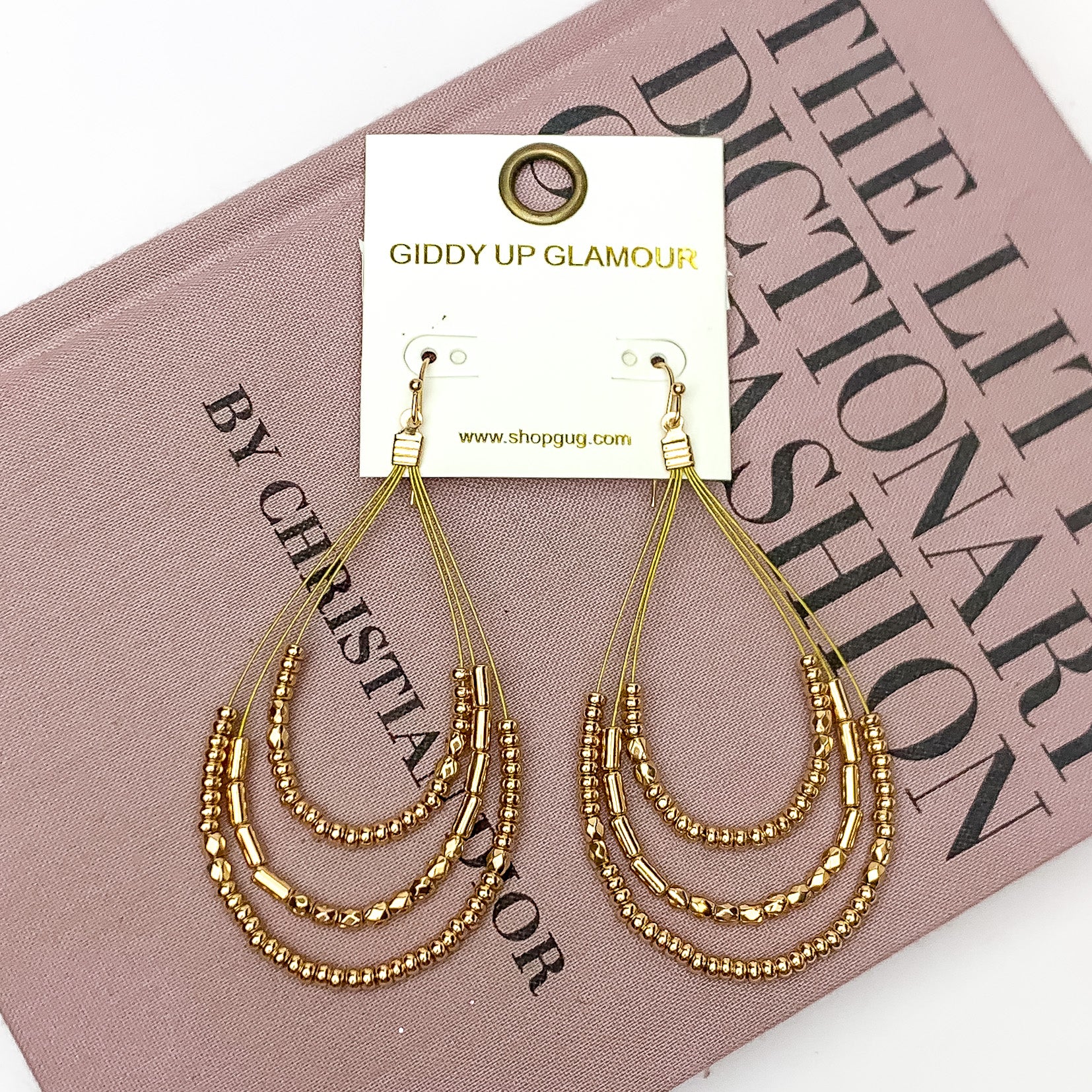 Layered Open Teardrop Earrings With Beads in Gold Tone. Pictured on a white background with the earrings laying on a pink book.