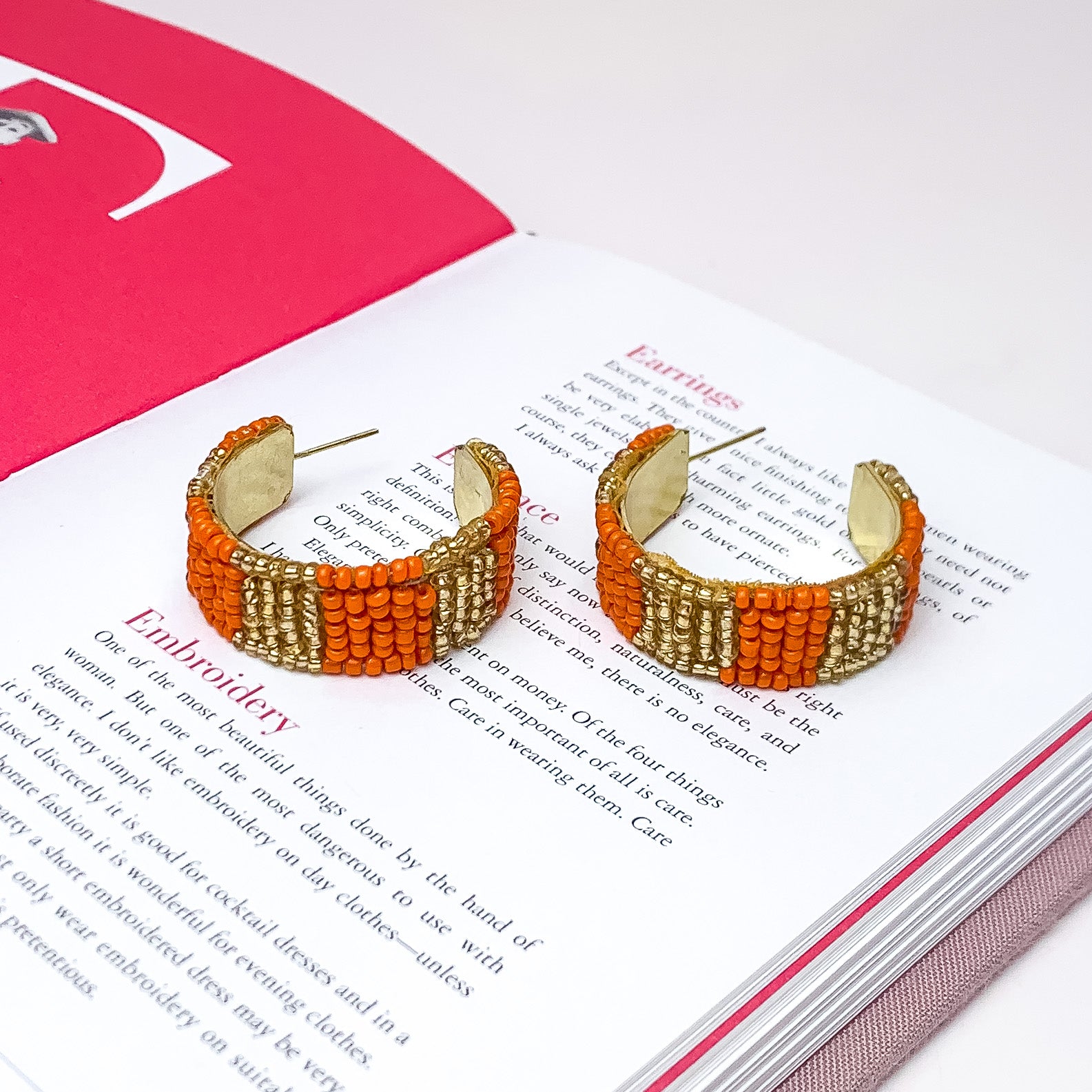 Vacay Era Gold Tone Beaded Hoop Earrings in Orange. Pictured on a white background with the earrings laying on an open book.