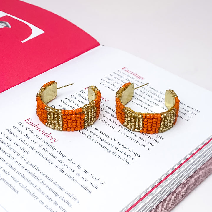 Vacay Era Gold Tone Beaded Hoop Earrings in Orange. Pictured on a white background with the earrings laying on an open book.
