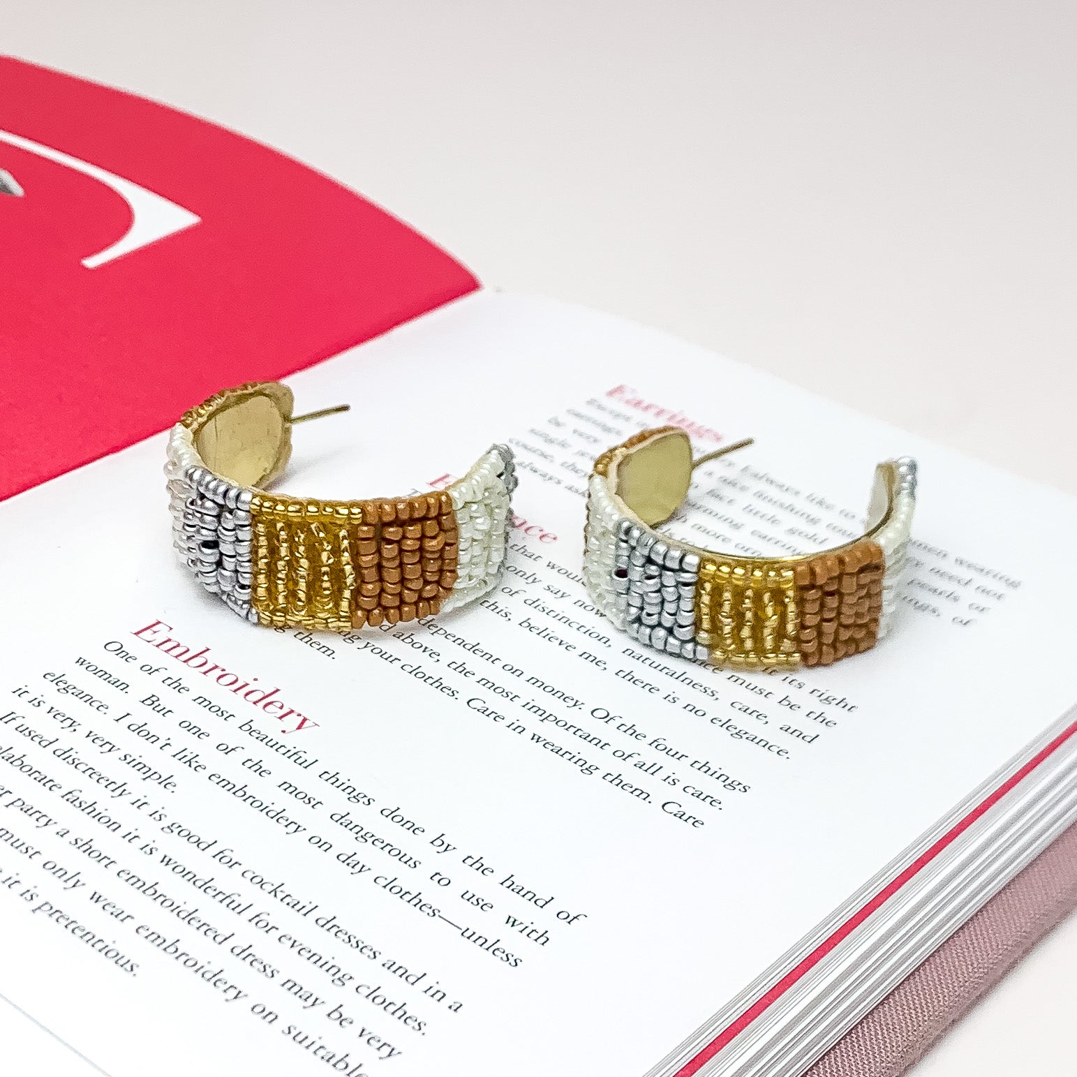 Vacay Era Gold Tone Beaded Hoop Earrings in Neutral Tones. Pictured on a white background with the earrings laying on an open page of a book.