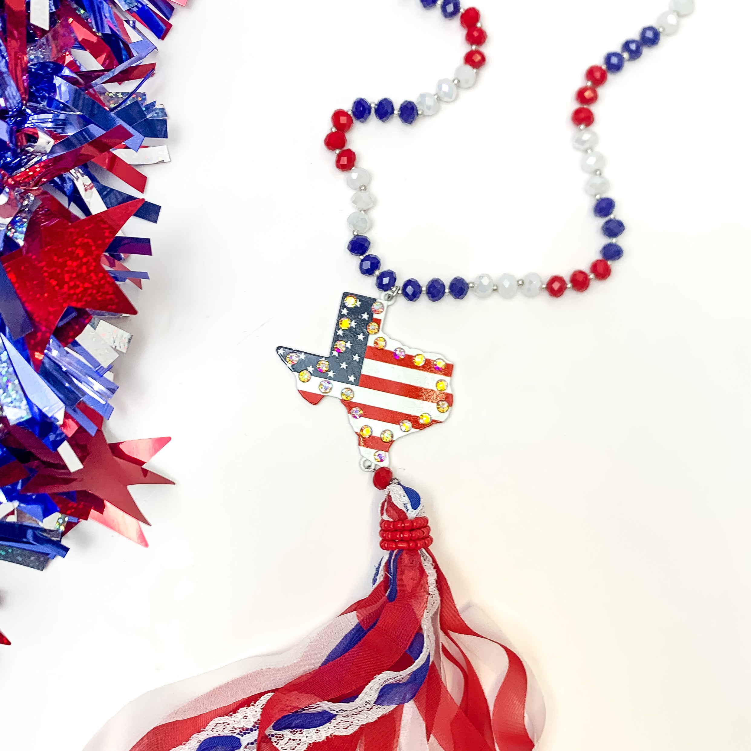 Red White and Bold USA Flag Texas Shape Necklace with Tassels. Pictured on a white background with red, white, and blue decoration to the left.