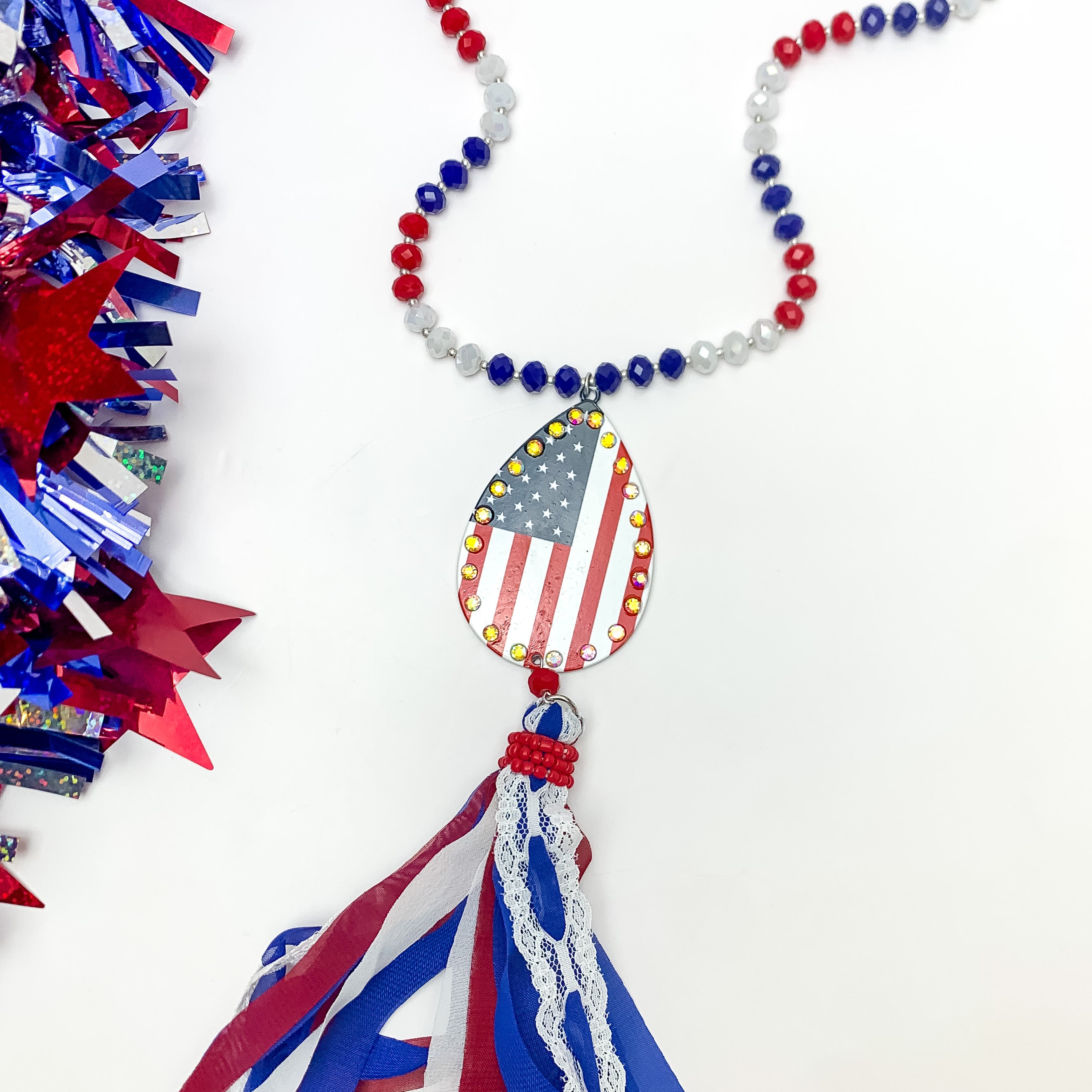 Red White and Bold USA Flag Necklace with Tassels. Pictured on a white background with red white and blue decoration on the left.