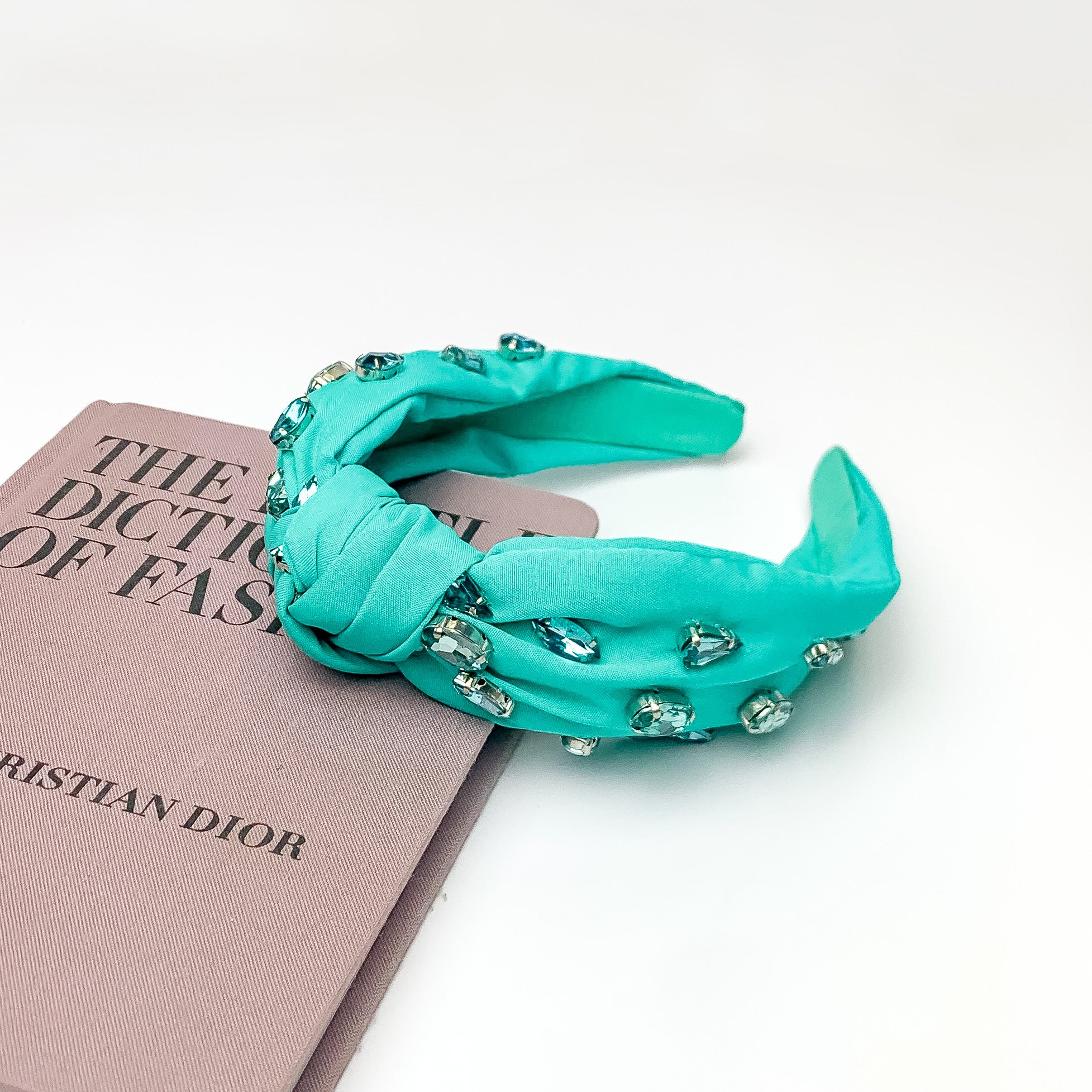 Crystal Detailed Knot Headband in Turquoise Green. Pictured on a white background with the headband laying against a pink book.