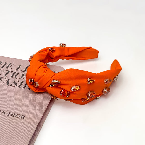 Crystal Detailed Knot Headband in Orange. Pictured on a white background with the headband laying against a pink book.