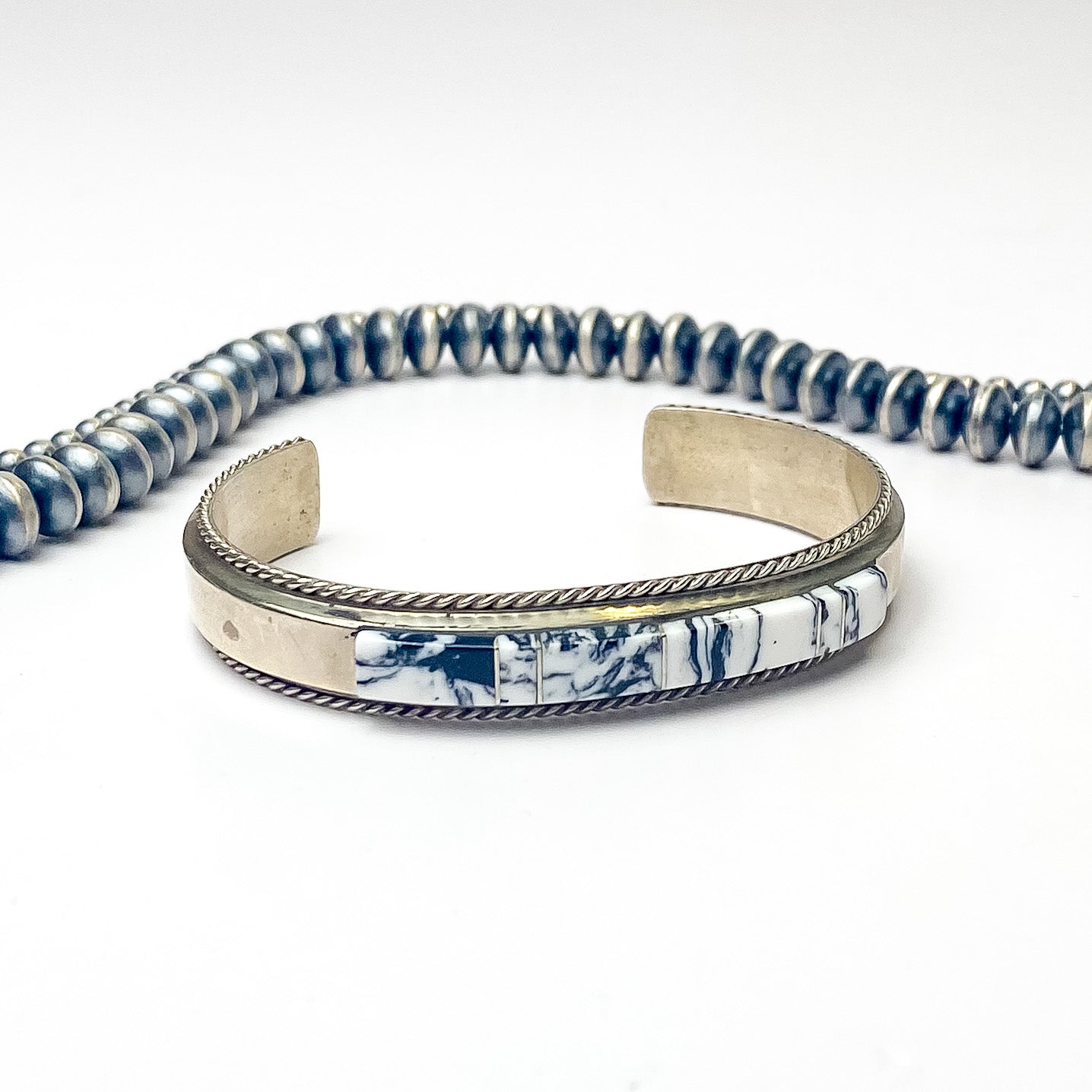Pictured is a silver bracelet with a rope detailing outline. This bracelet also includes white buffalo stones. This bracelet is pitured on a white background with silver beads above the bracelet. 