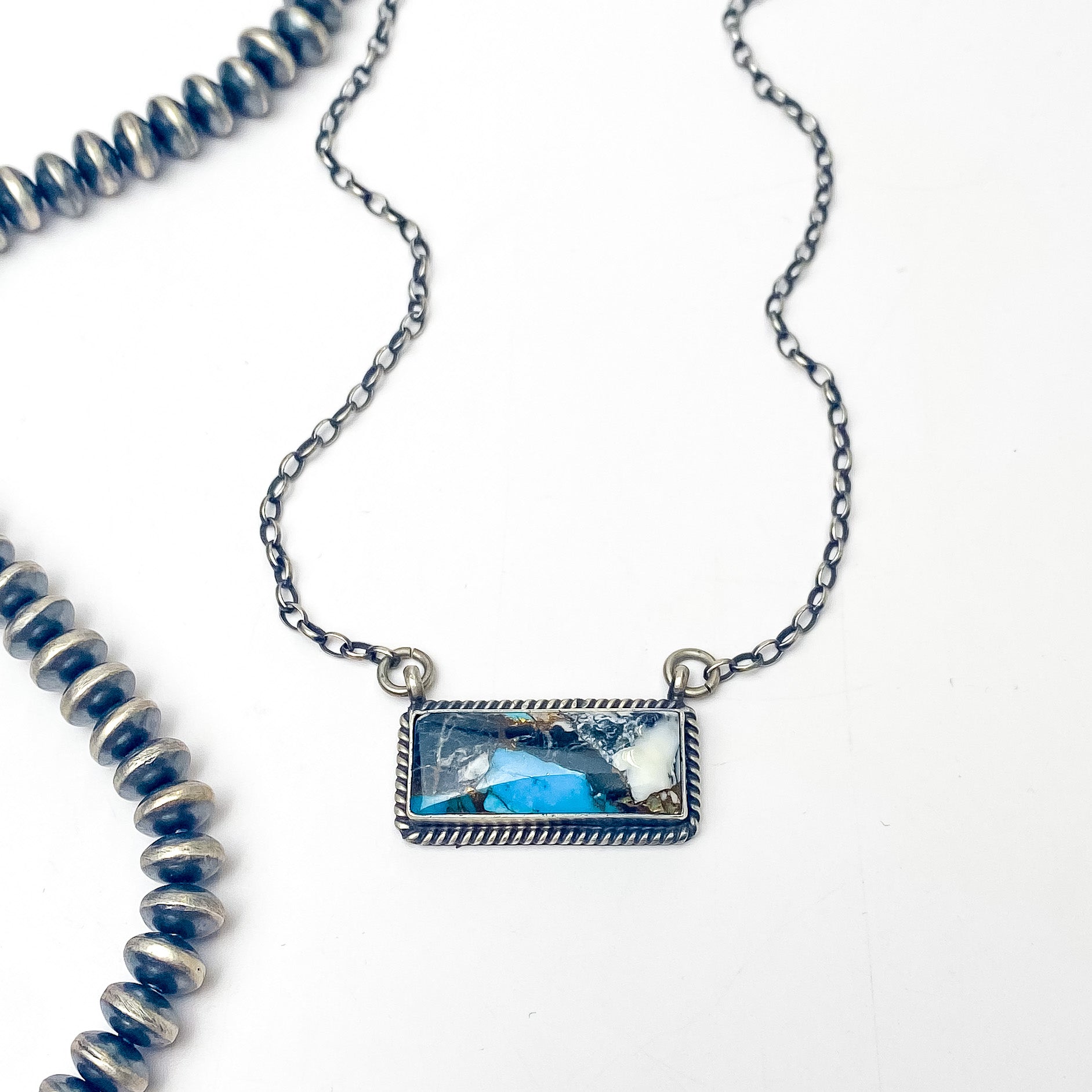Pictured is a silver chain necklace with a silver bar pendant and a white buffalo and turquoise remix stone. This necklace is pictured on a white background with silver beads on the left side. 