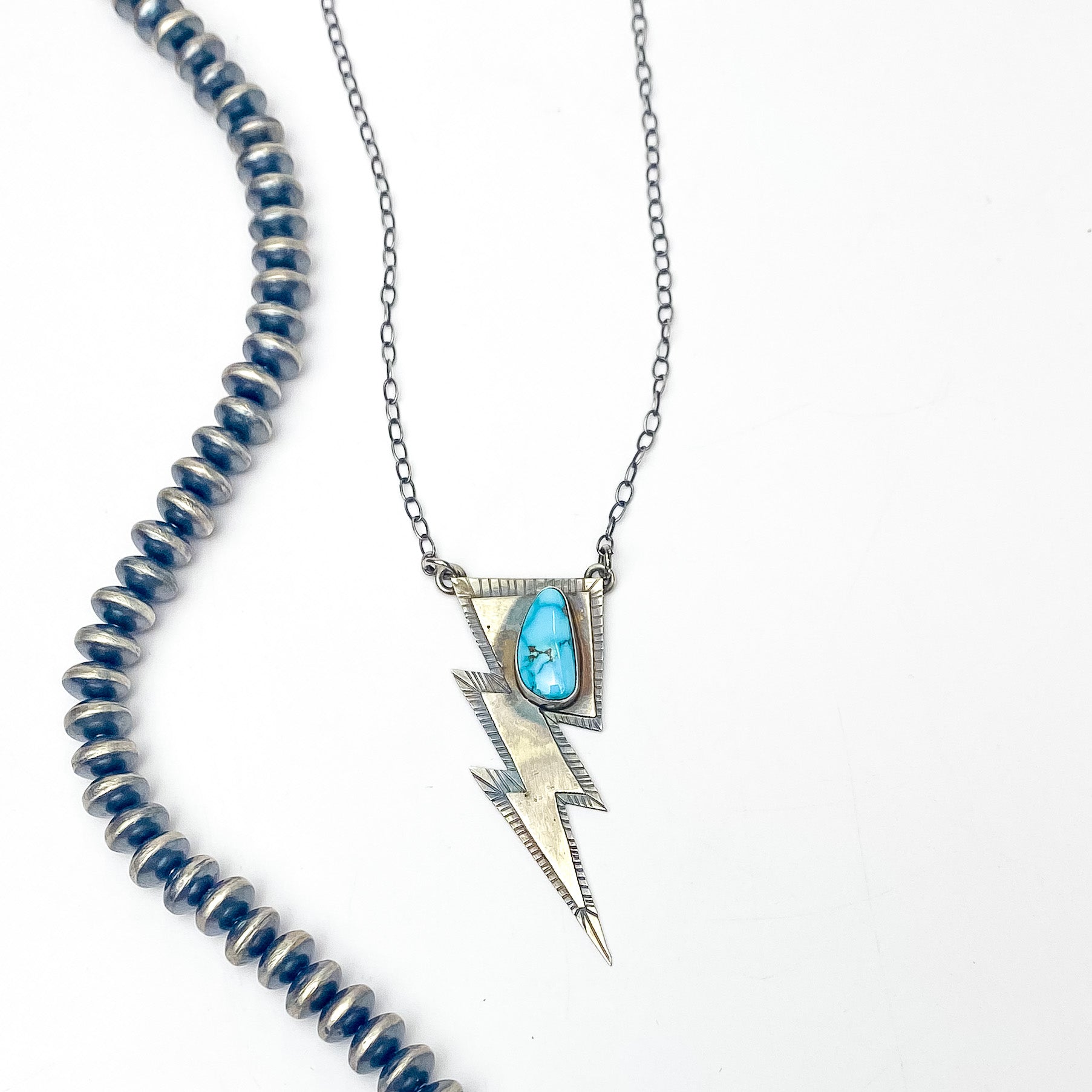 Pictured is a silver chain necklace with a silver lightning bolt pendant and a turquoise stone. This necklace is pictured on a white background with silver beads on the left side. 