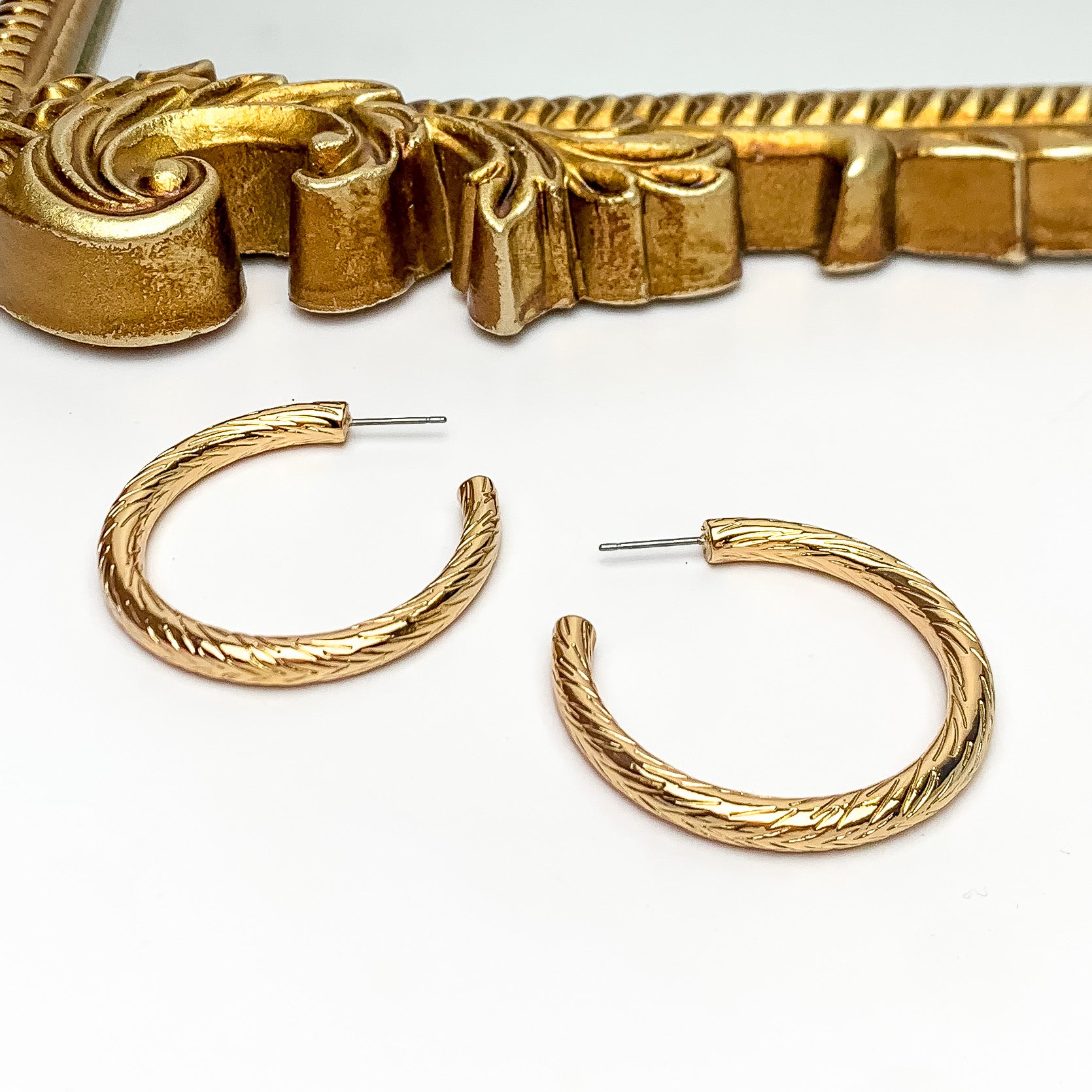 Gold Tone Large Twisted Hoop Earrings. Pictured on a white background with a gold frame above the earrings.