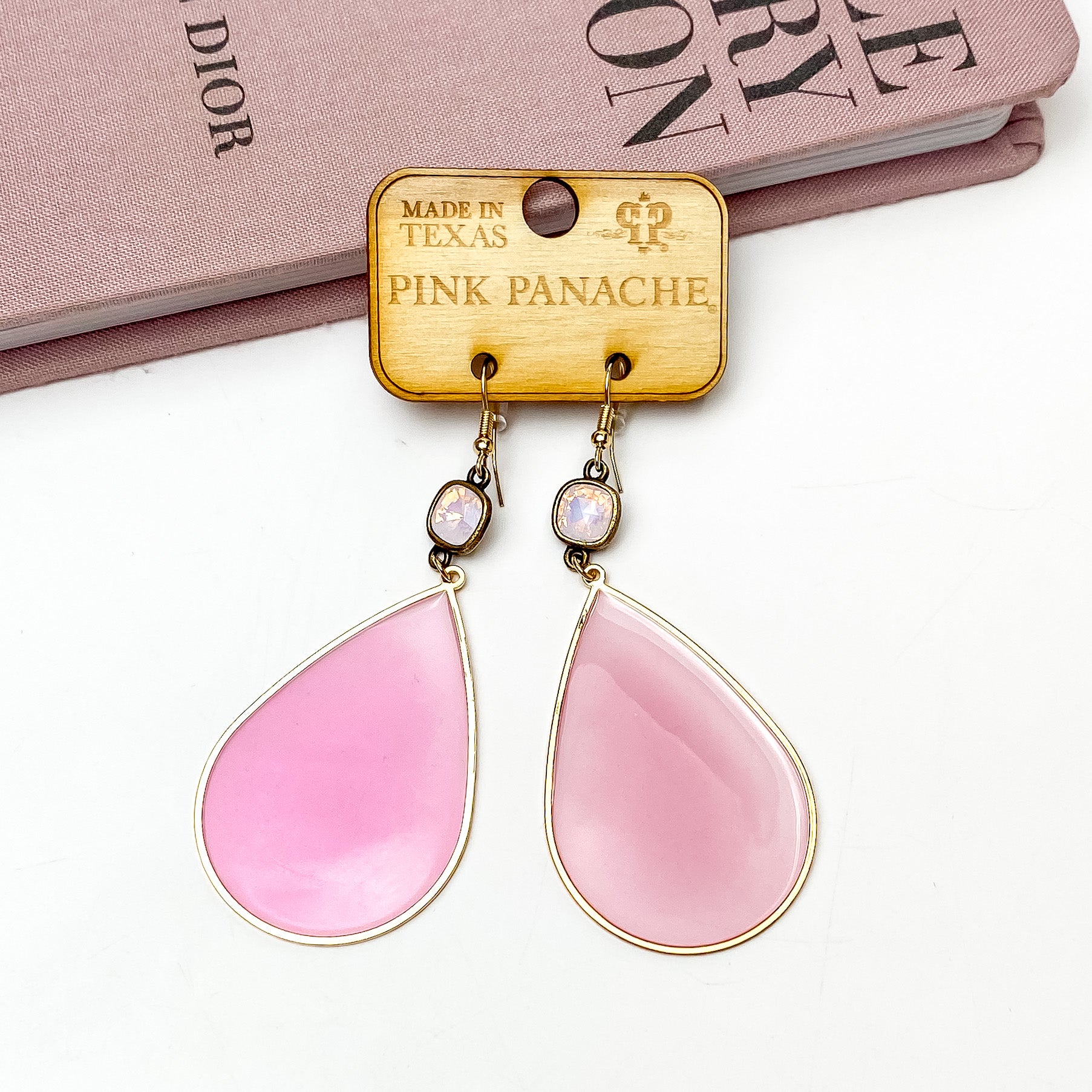 Rose water opal cushion cut crystal drop earrings a pink acrylic teardrop pendant outlined in gold. These earrings are pictured on a wooden earring holder on a white background in front of a mauve colored book. 