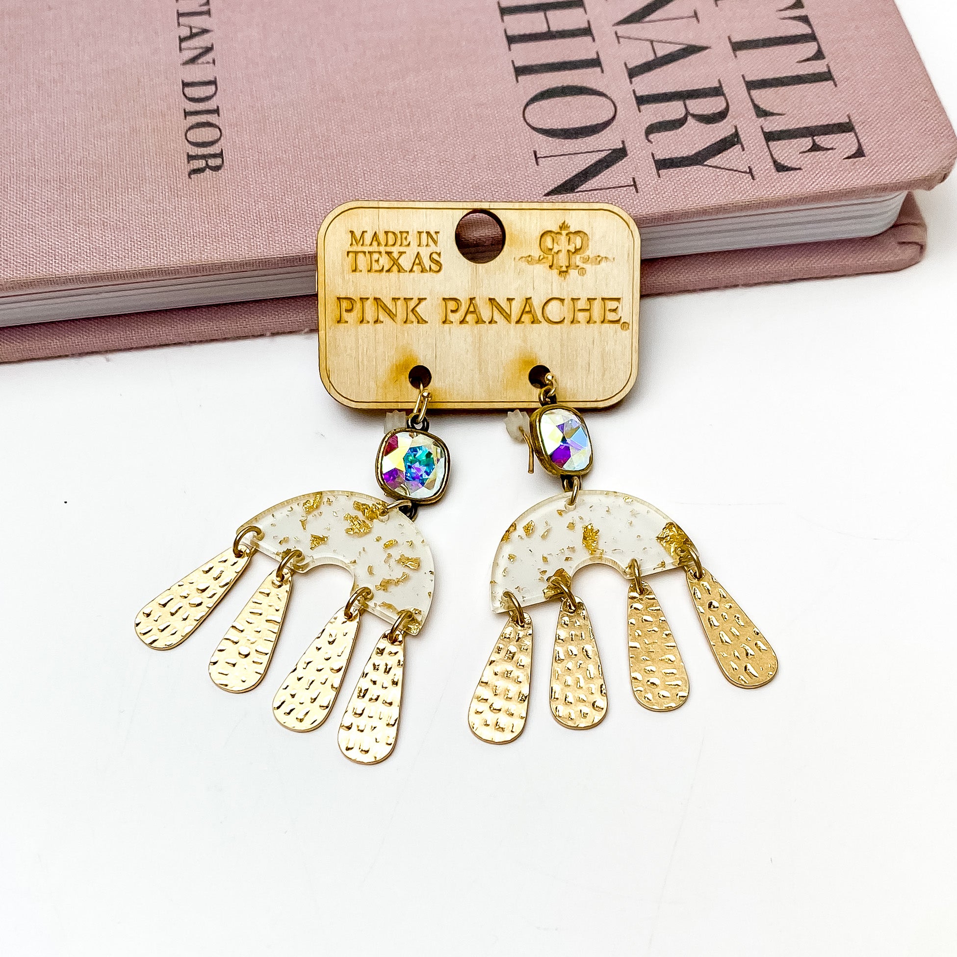 AB cushion cut crystal drop earrings with a half circle, white and gold pendant with gold raindrop charms. These earrings are pictured on a wooden earring holder on a white background in front of a mauve colored book. 