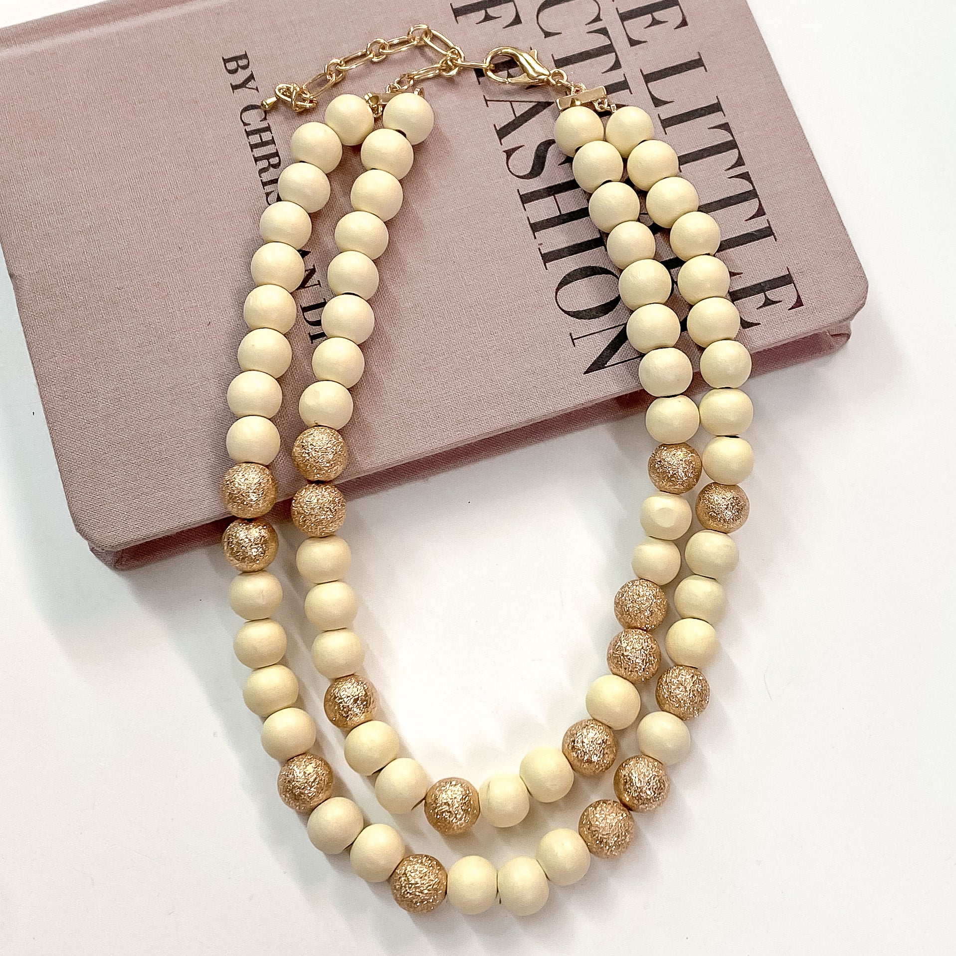 Pictured partially on a mauve colored book on a white background is a two strand ivory beaded necklace with gold beaded spacers and gold hardware. 