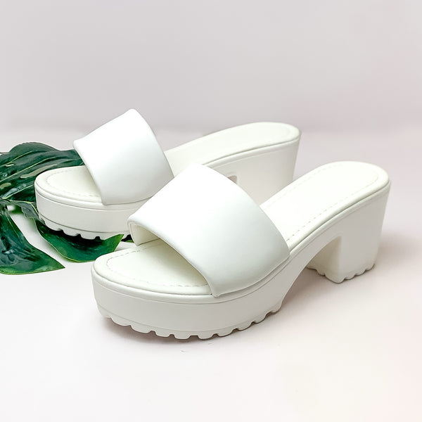 Pictured is a piar of platform, side on sandals with a block heel and one strap in white. These shoes are pictured on a white background with a green leaf under on of the shoes. 