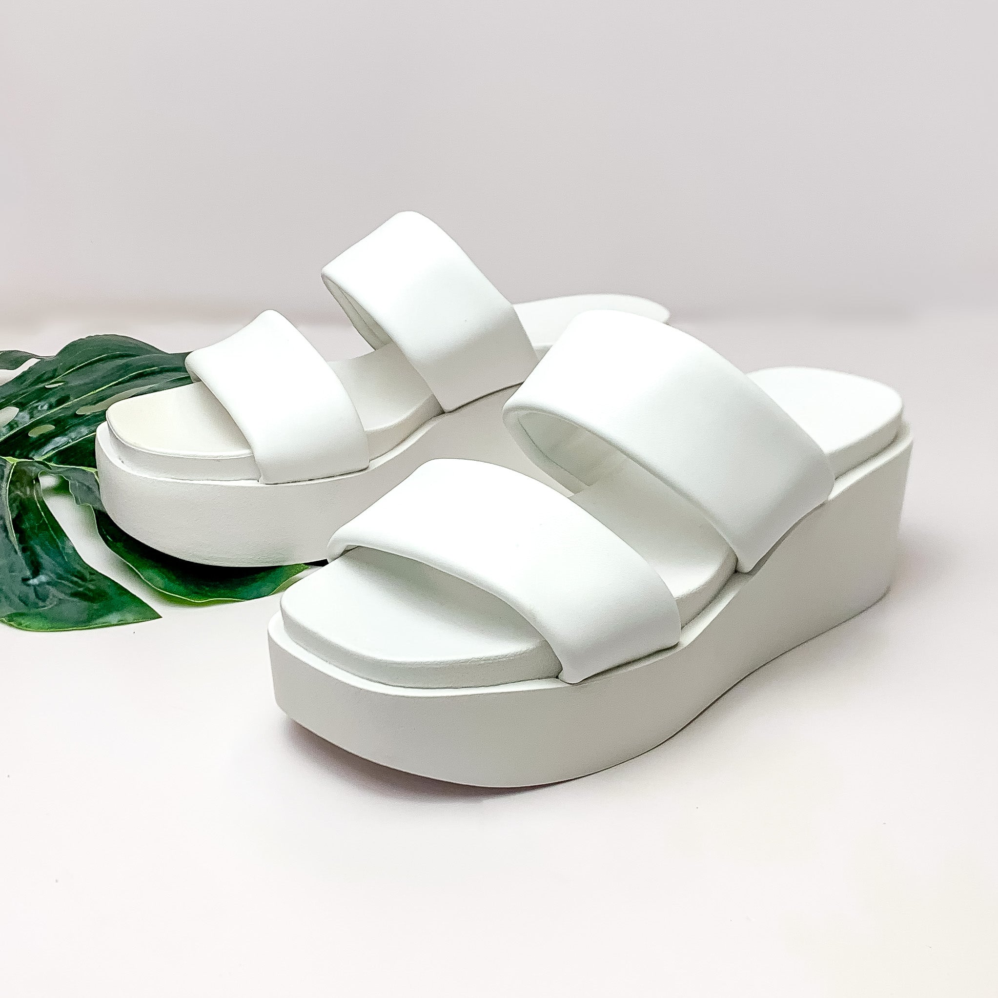 Pictured is a pair of platform, side on sandals with a 2 straps in white. These shoes are pictured on a white background with a green leaf under one of the shoes. 