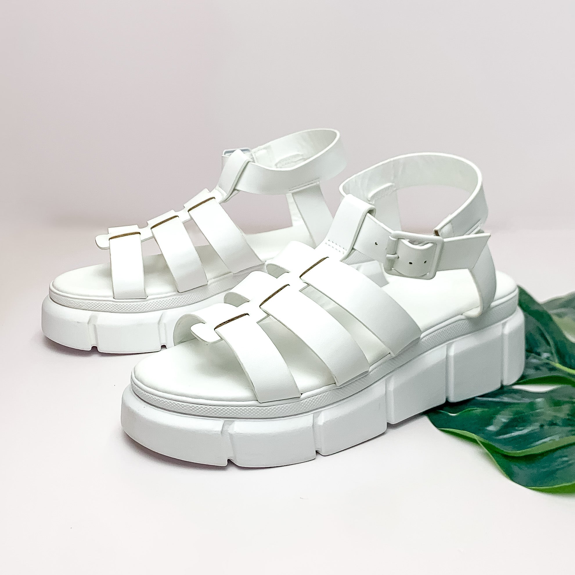 Pictured is a pair of platform, gladiator sandals with an adjustable ankle strap. These shoes are pictured on a white background with a green leaf under one of the shoes. 