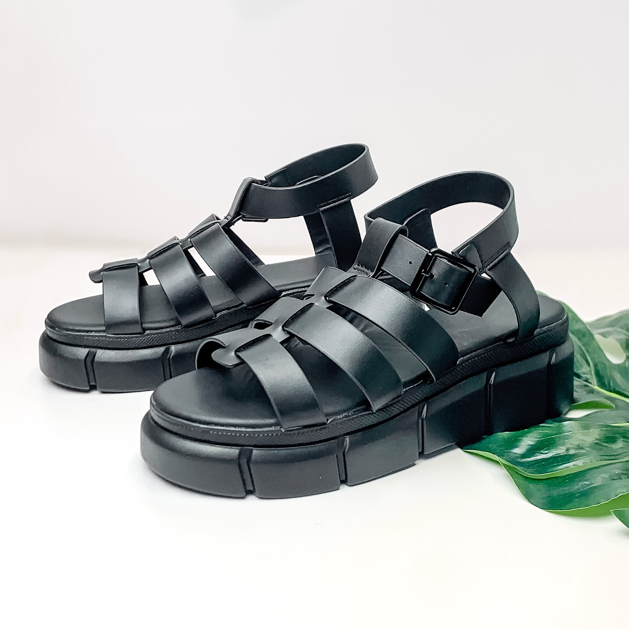 Pictured is a pair of platform, gladiator sandals with an adjustable ankle strap in black. These shoes are pictured on a white background with a green leaf under one of the shoes. 