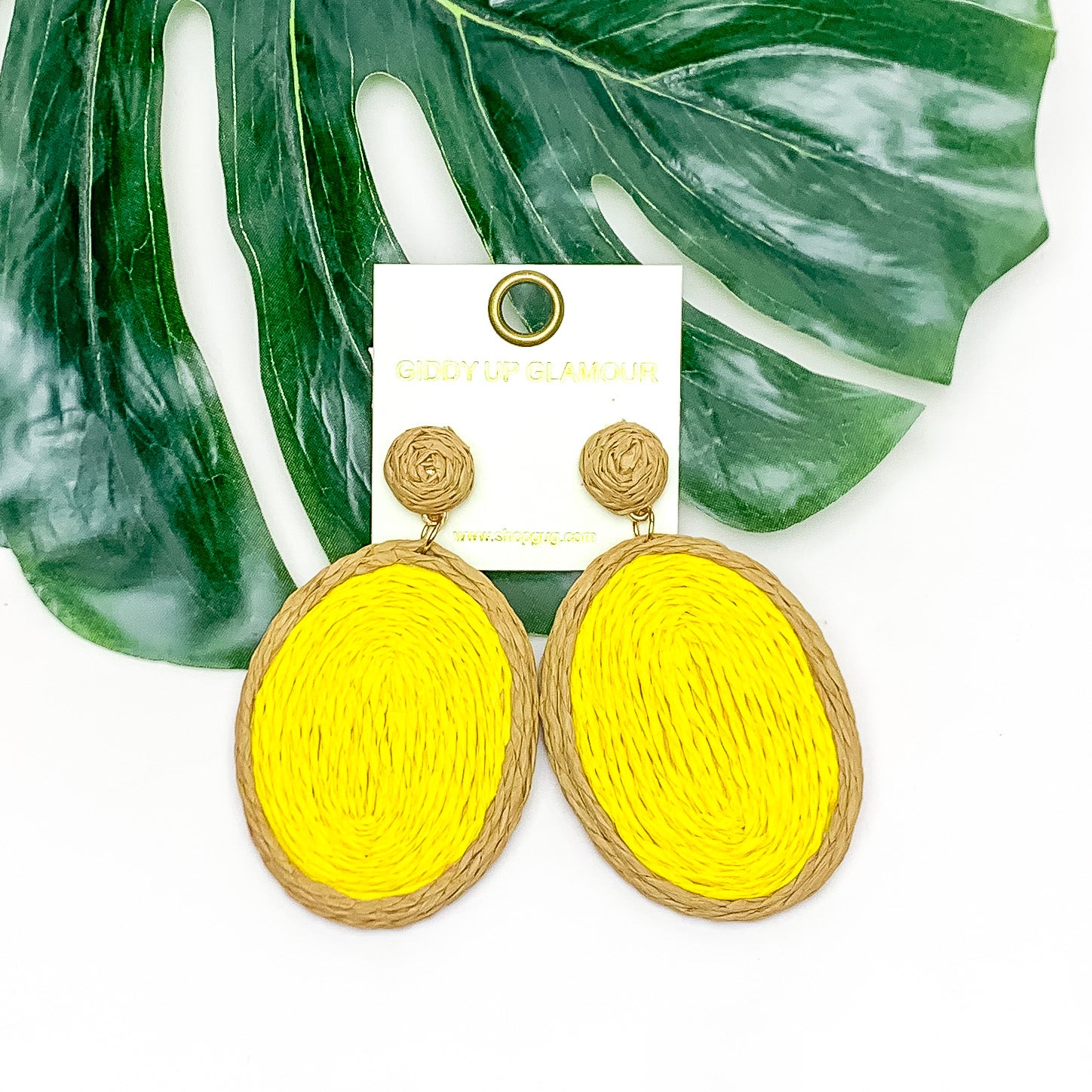 Brunch Bash Raffia Wrapped Oval Earrings in Yellow. Pictured on a white background with a large leaf behind the earrings.