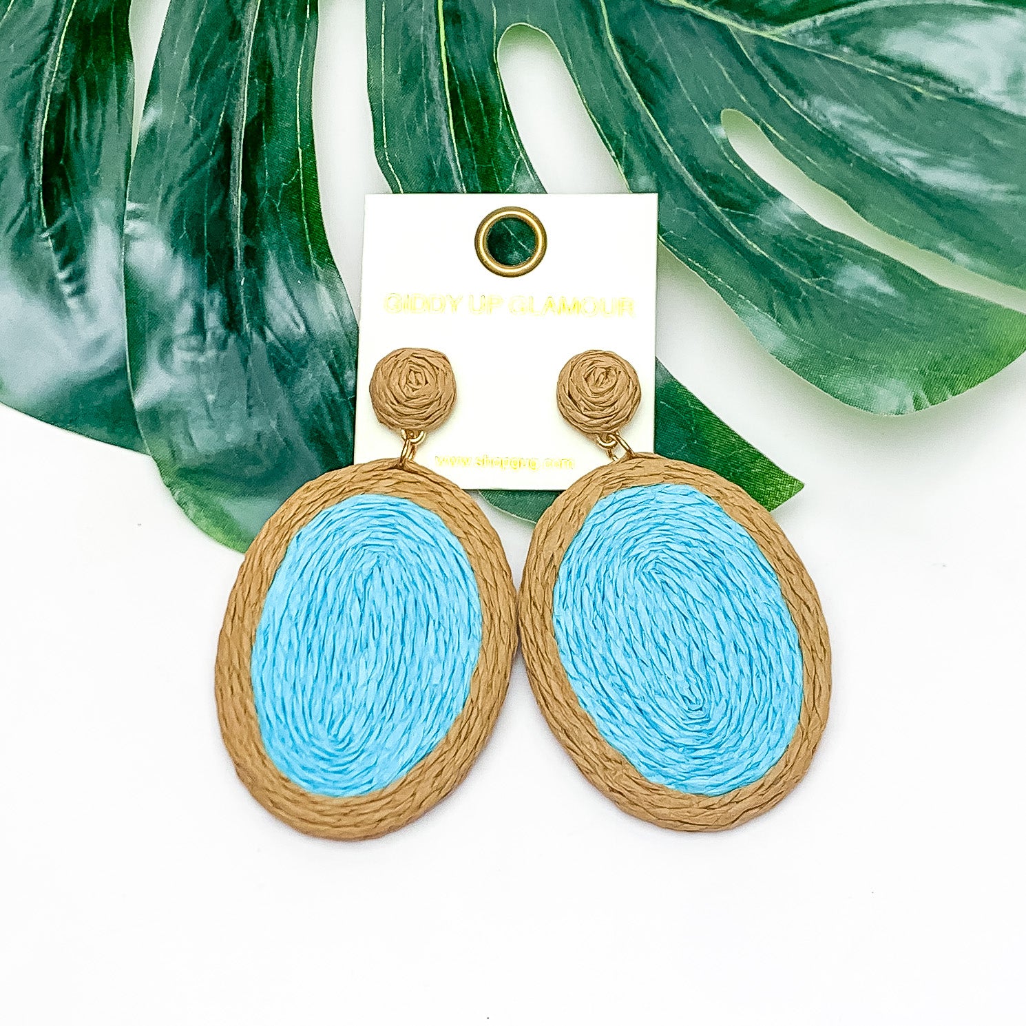 Brunch Bash Raffia Wrapped Oval Earrings in Light Blue. Pictured on a white background with a large leaf behind the earrings.