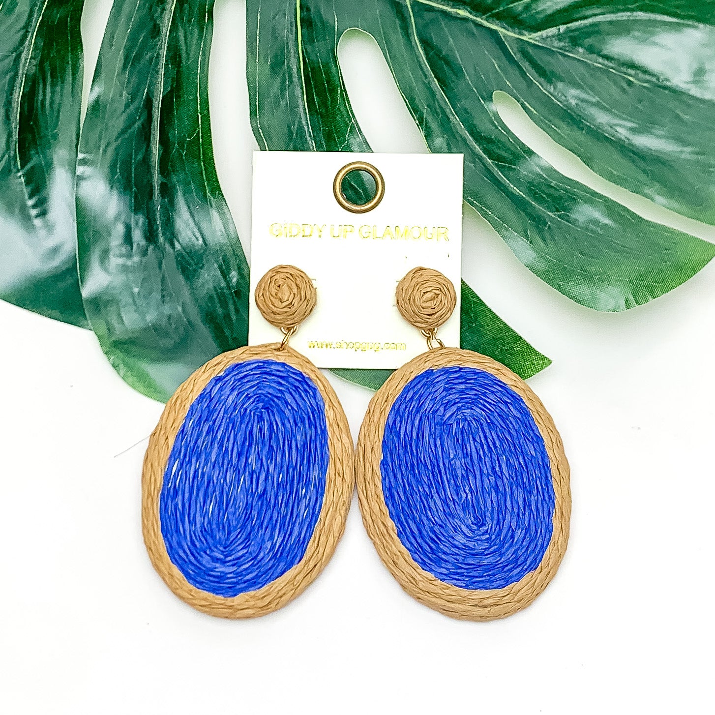 Brunch Bash Raffia Wrapped Oval Earrings Royal Blue. Pictured on a white background with a large leaf behind the earrings.