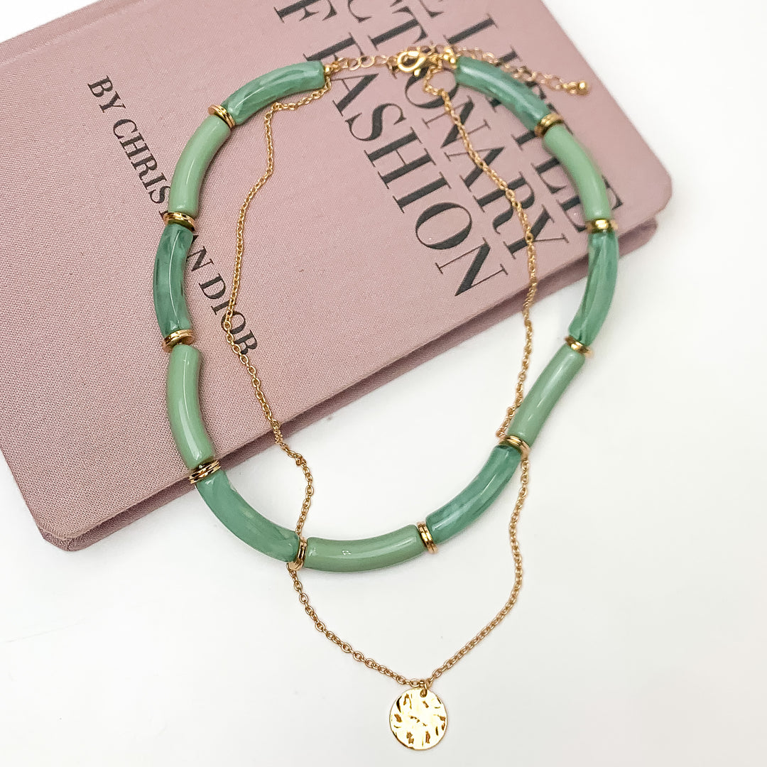 Perfect Paradise Tube Necklace With Second Gold Tone Chain Necklace in Greene. Pictured on a white background with the necklace laying on a book.