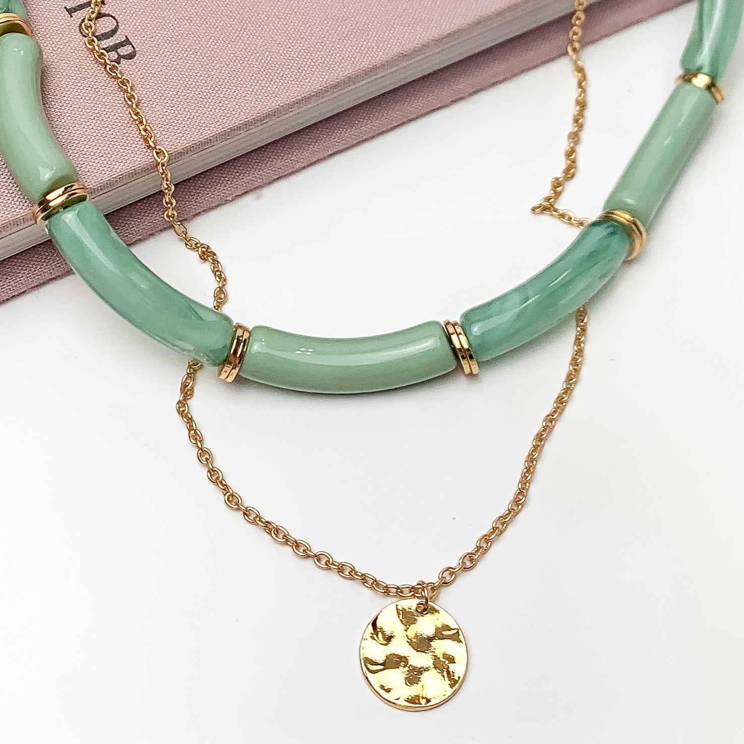 Perfect Paradise Tube Necklace With Second Gold Tone Chain Necklace in Greene. Pictured on a white background with the necklace laying on a book.