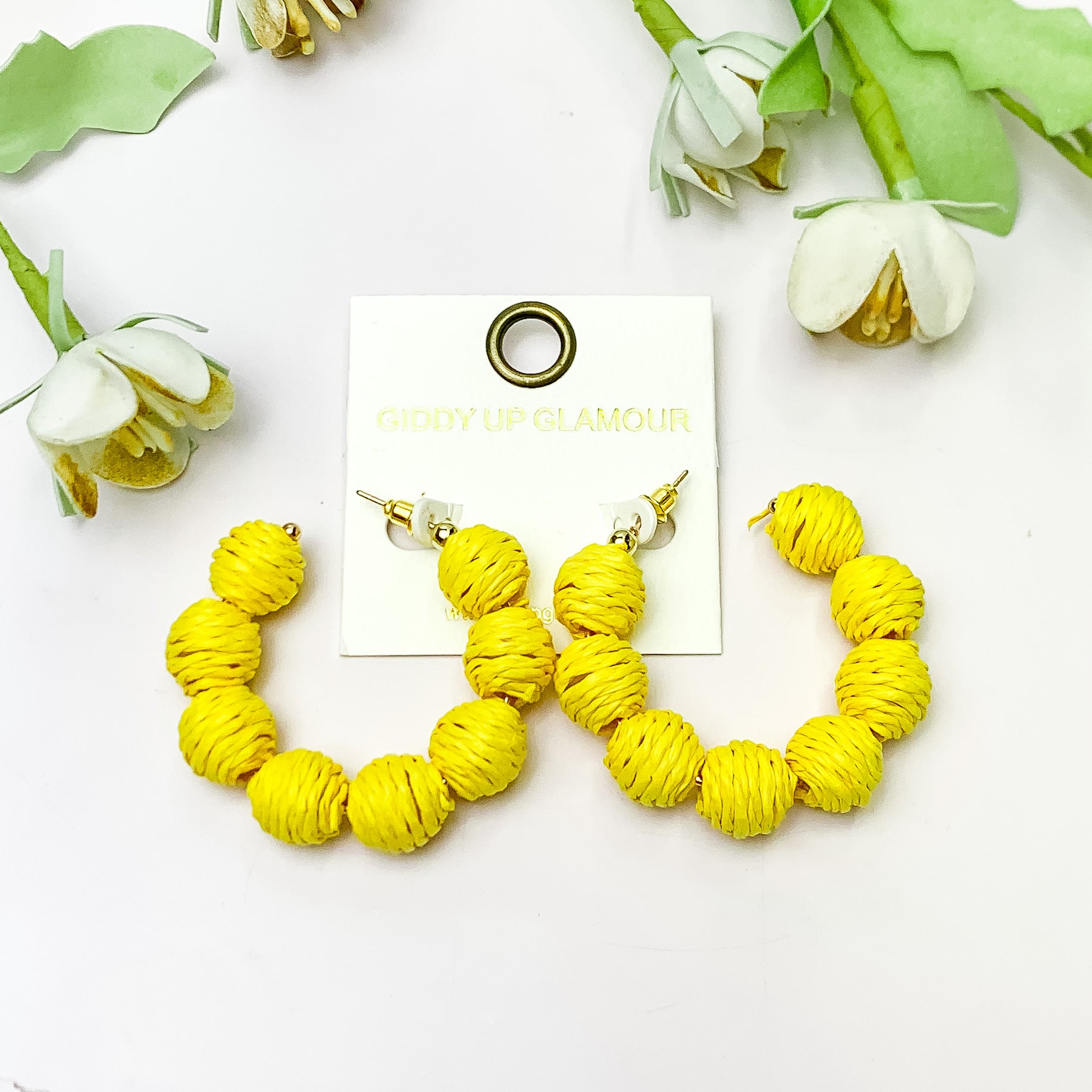 Sorbet Summer Raffia Ball Hoop Earrings in Yellow. Pictured on a white background with flowers above the earrings.