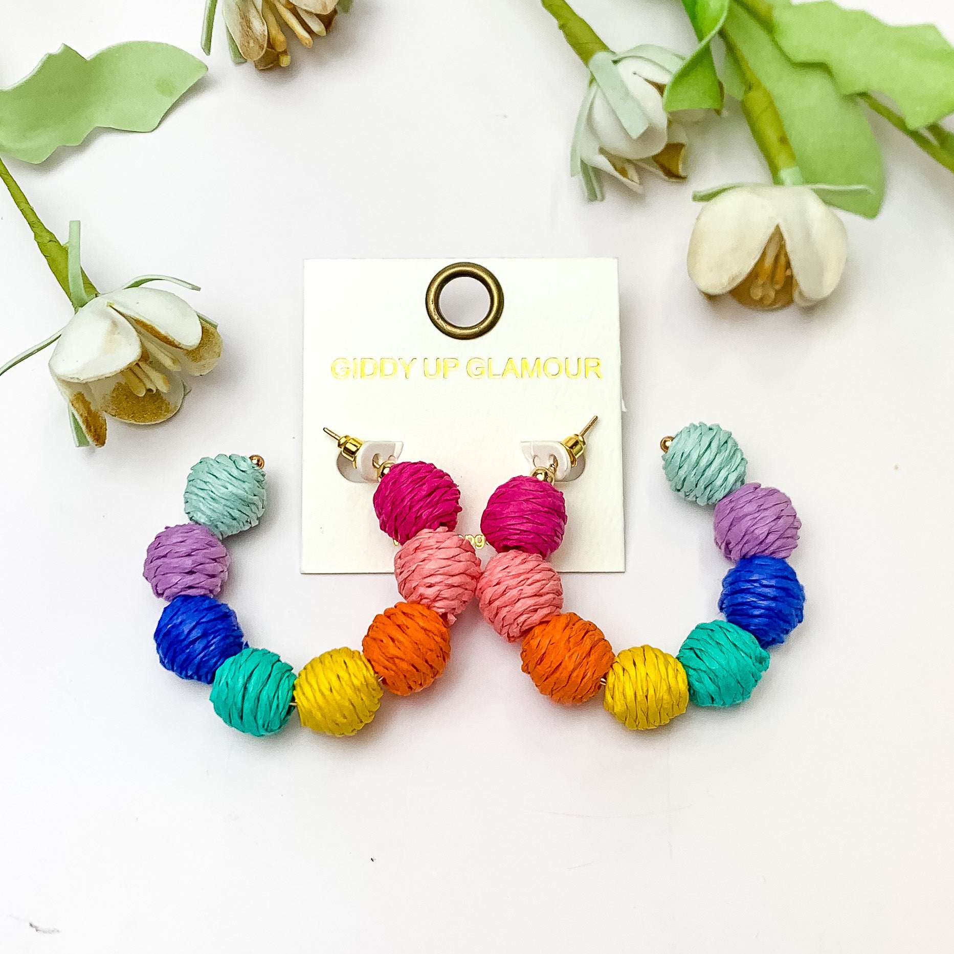 Sorbet Summer Raffia Ball Hoop Earrings in Multicolor. Pictured on a white background with flowers laying above the earrings.