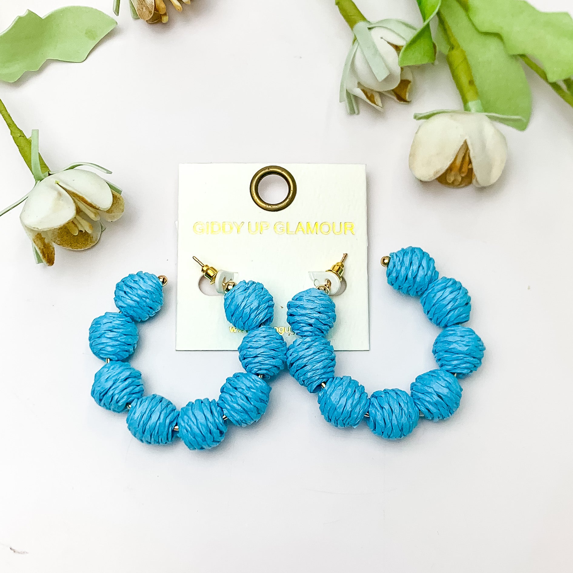 Sorbet Summer Raffia Ball Hoop Earrings in Blue. Pictured on a white background with flowers above the earrings.