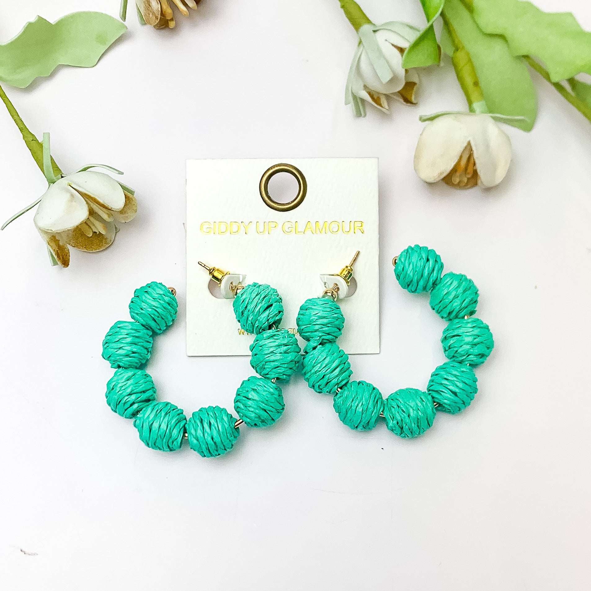 Sorbet Summer Raffia Ball Hoop Earrings in Turquoise Green. Pictured on a white background with flowers above the earrings.