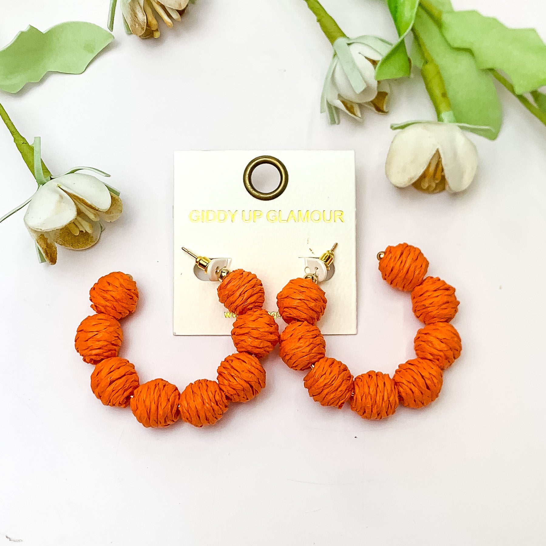 Sorbet Summer Raffia Ball Hoop Earrings in Orange. Pictured on a white background with flowers above the earrings. 