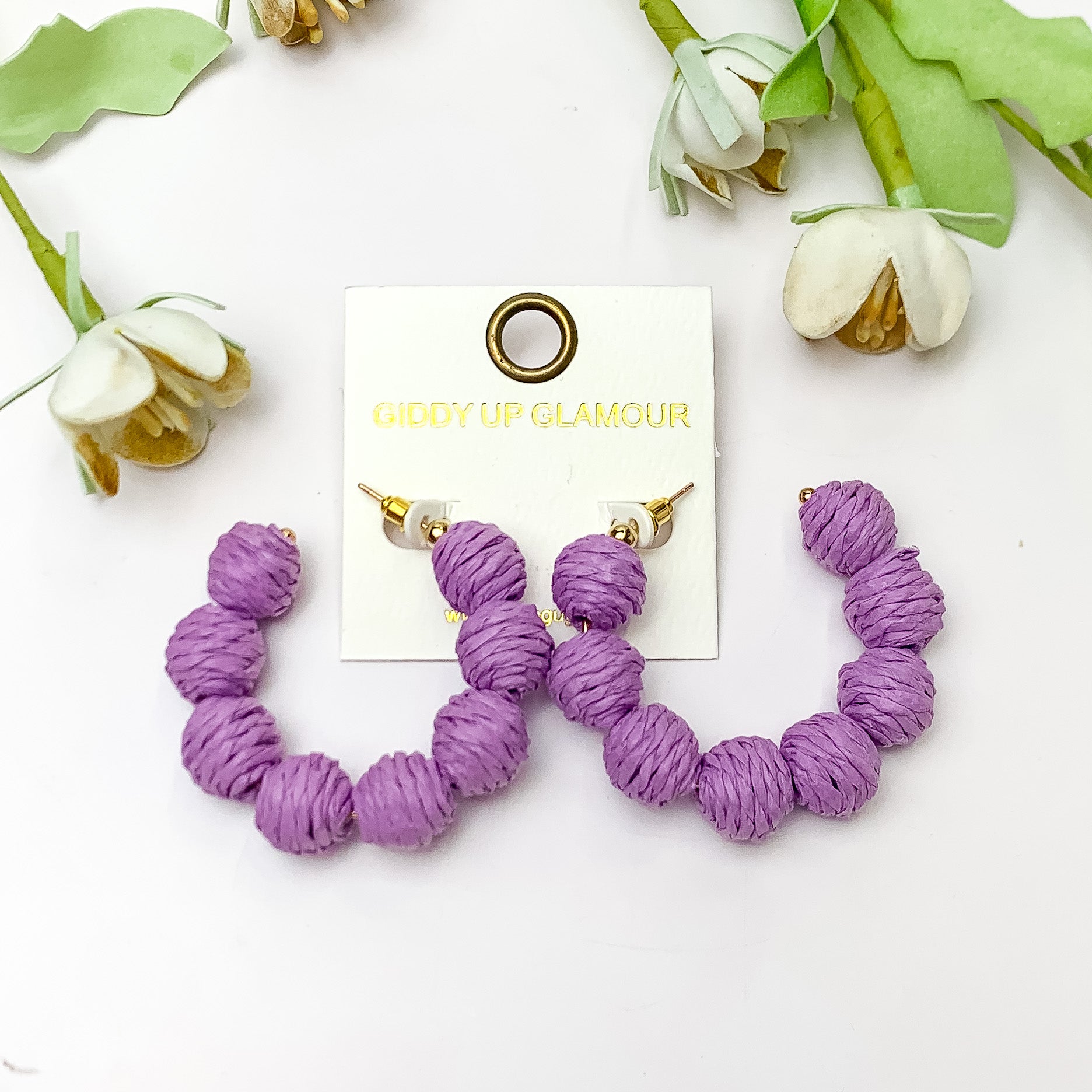 Sorbet Summer Raffia Ball Hoop Earrings in Lavender Purple. Pictured on a white background with flowers above the earrings.